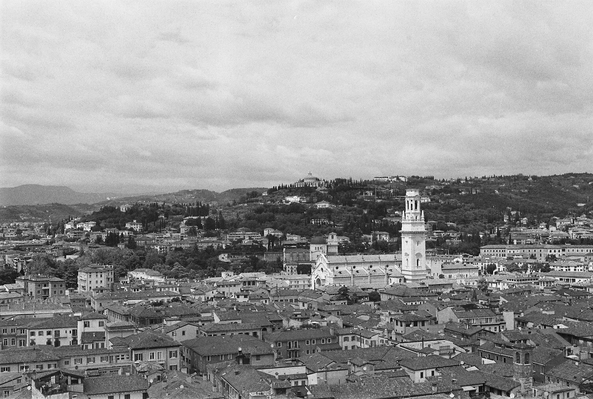 Black-and-white elevated cityscape of Verona with tiled roofs, a square bell tower and wooded hills beyond.