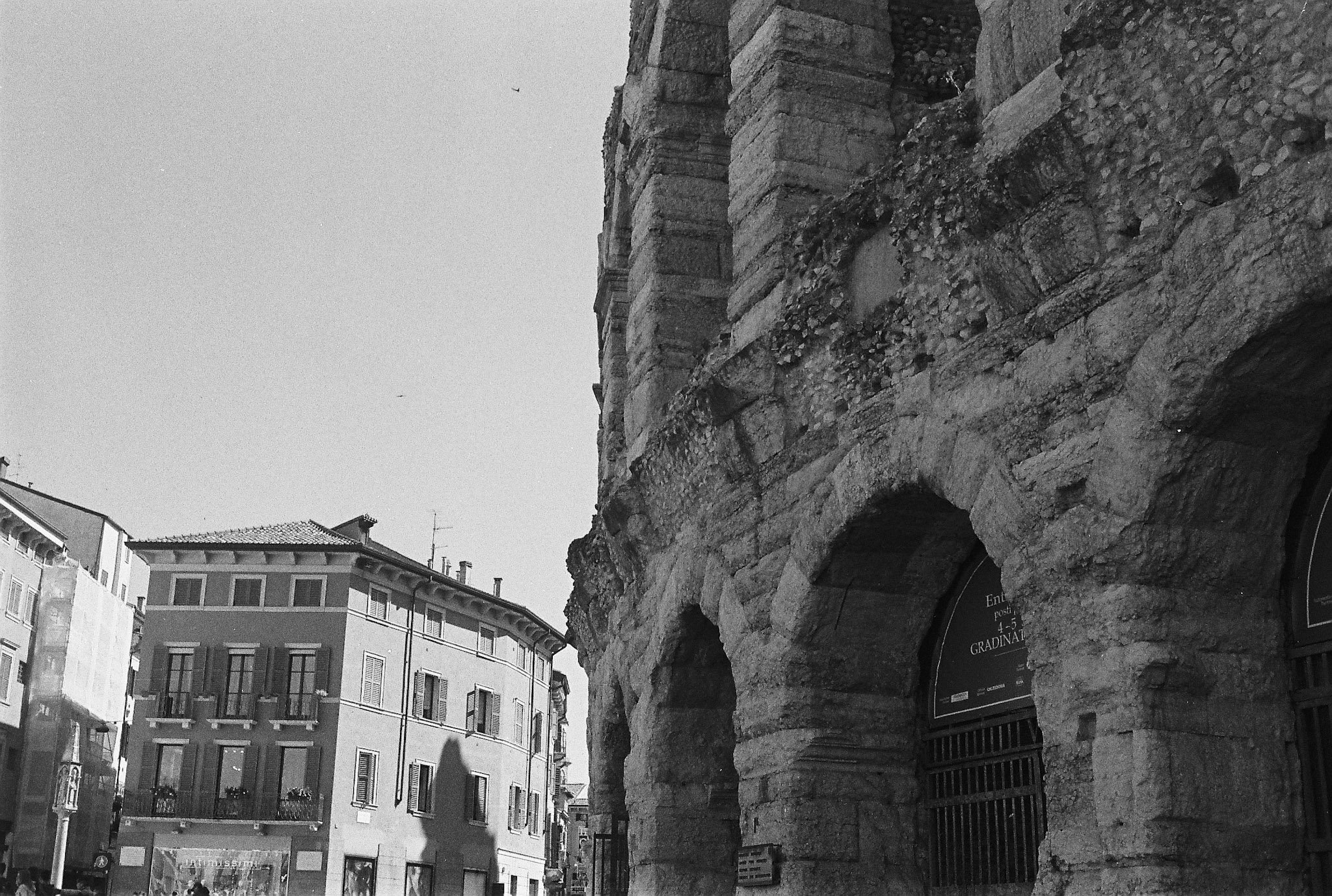 Black-and-white street view of a large weathered stone structure beside stucco buildings in Verona.