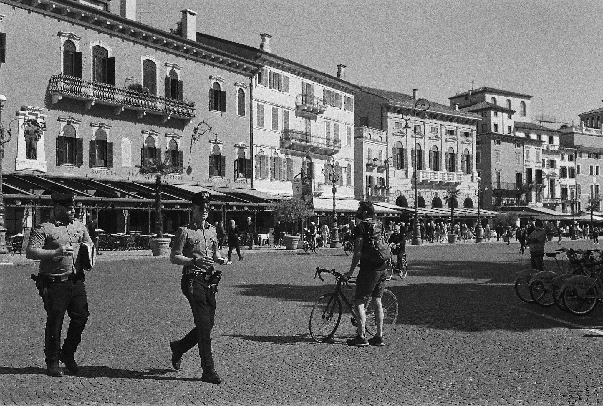 Black-and-white piazza scene in Verona with bicycles, two police officers, historic facades and café awnings.