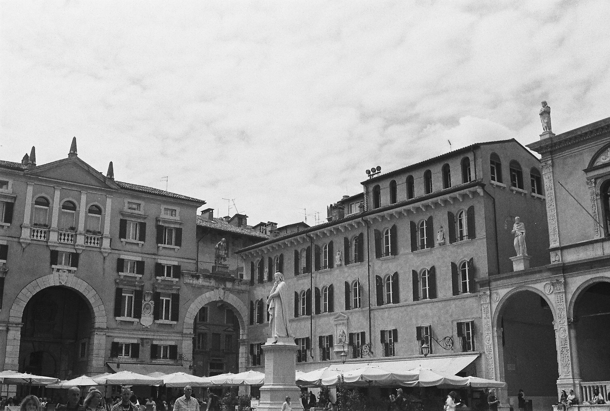 Black-and-white photograph of a historic Verona square with arcaded buildings, statues, umbrellas and pedestrians.