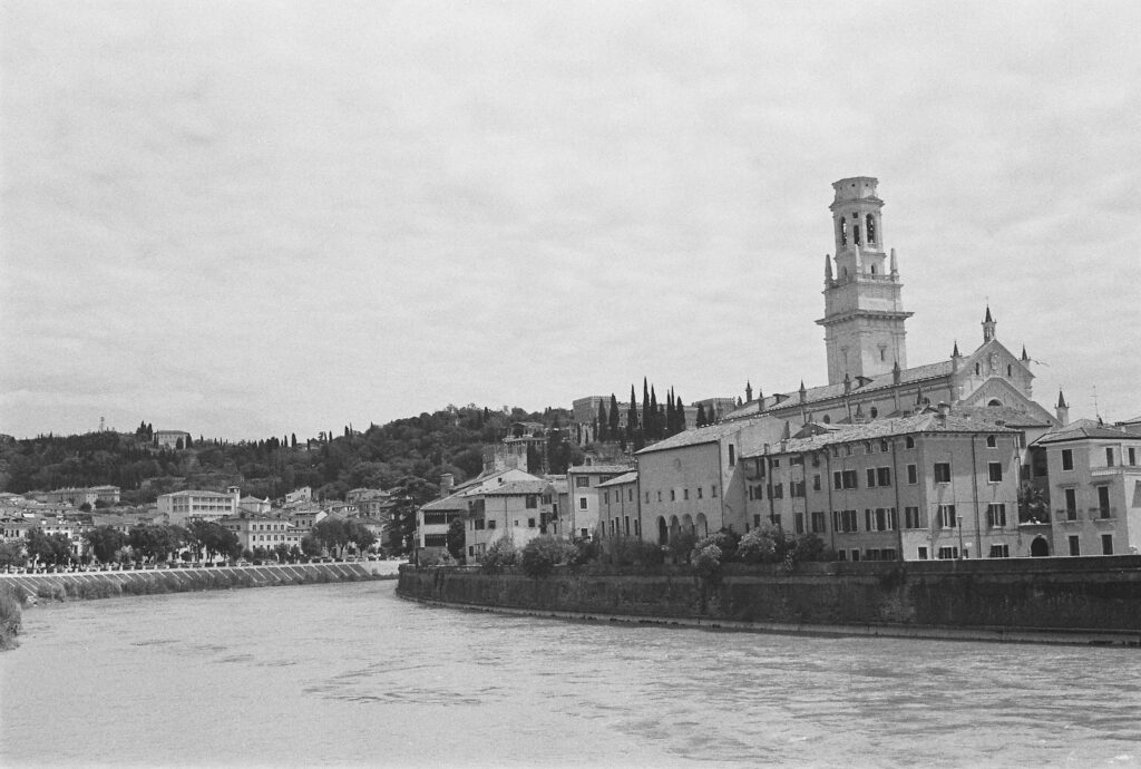 Black-and-white view of Verona’s riverfront with embankments, dense historic buildings, a bell tower and hillside cypress trees.