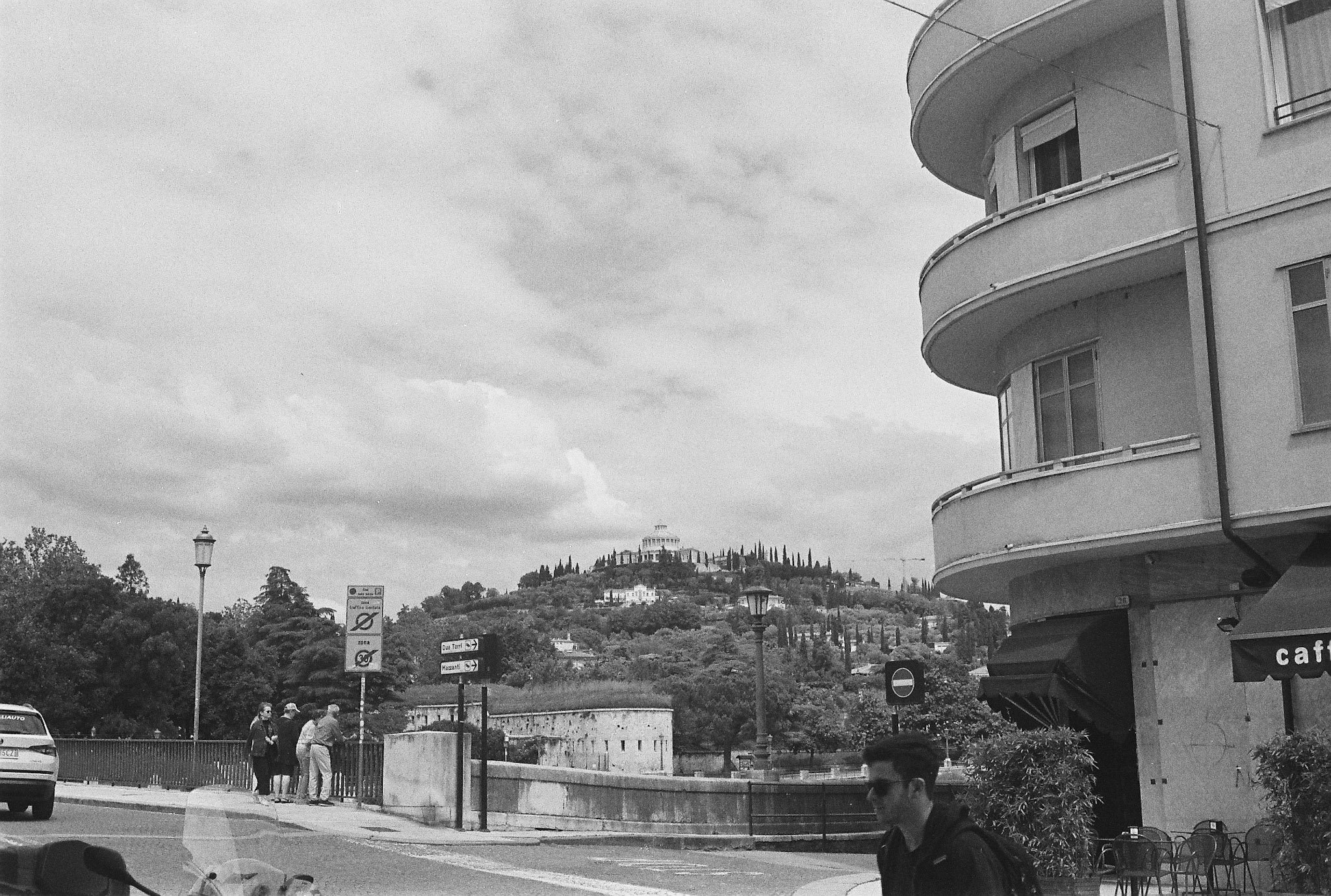 Black-and-white Verona street scene with traffic-limited signage, pedestrians, curved balconies and a landscaped hill in the background.