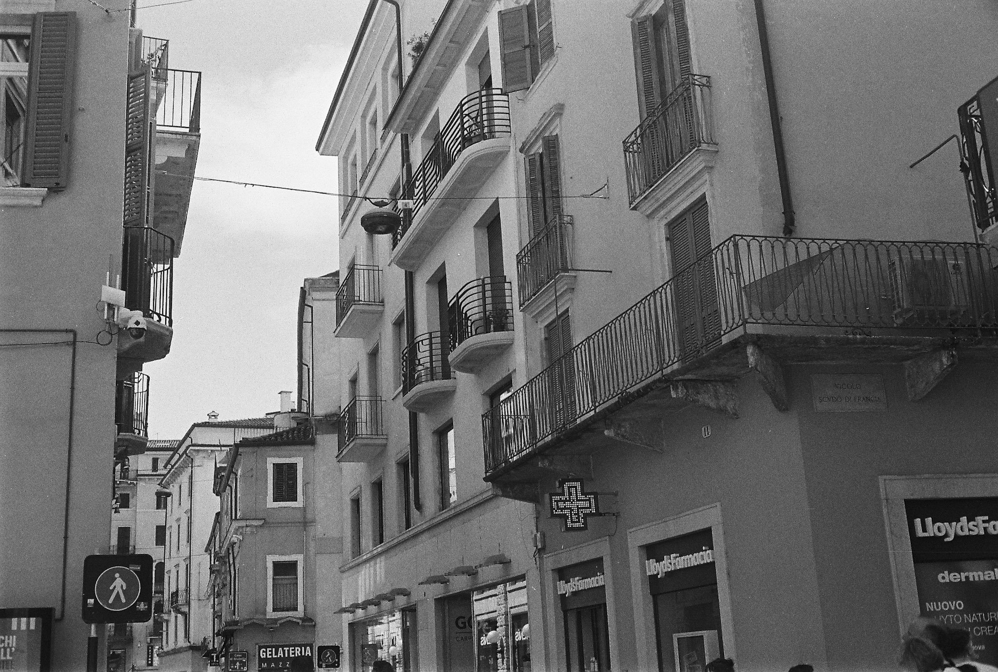 Black-and-white narrow Verona street with balconies, storefronts, Lloyds pharmacy sign and Gelateria Mazzi sign.