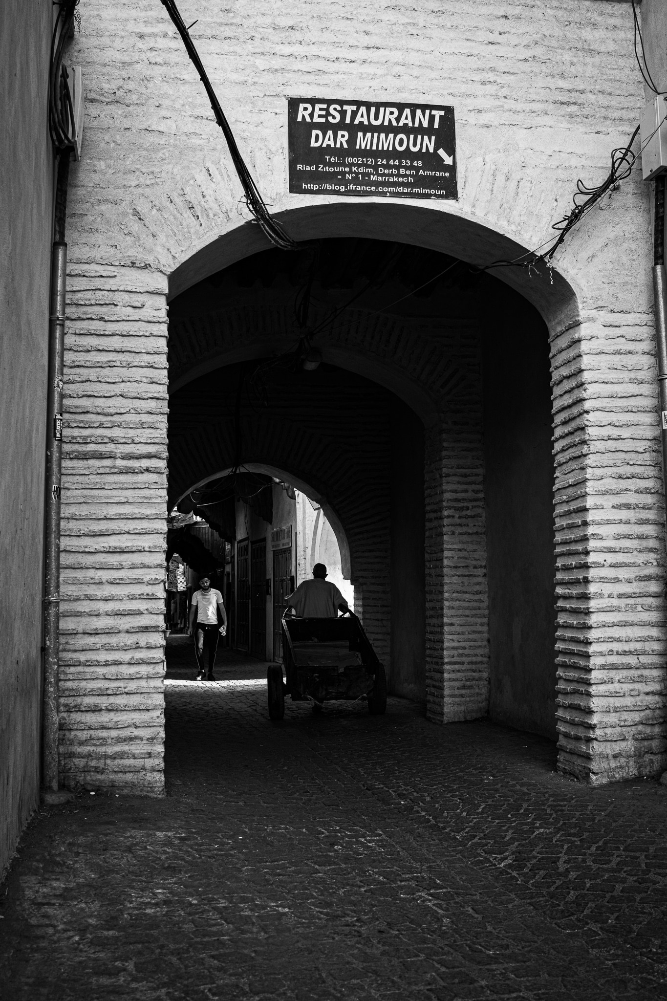 Arched medina passage in Marrakech with pedestrians and a handcart beneath restaurant signage