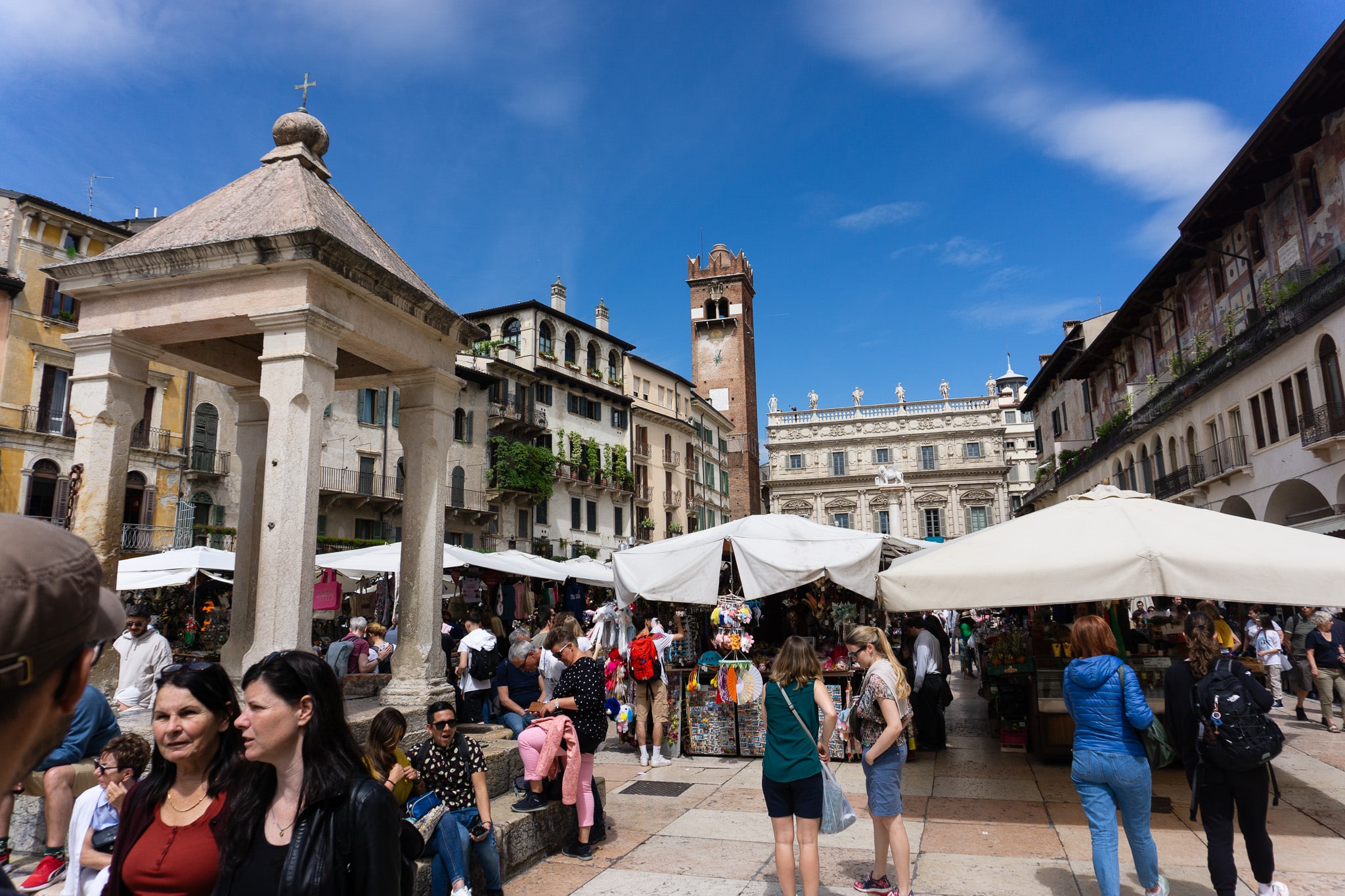 Open-air market in Verona’s historic square with white umbrellas, crowds, frescoed buildings, and a tall brick tower.