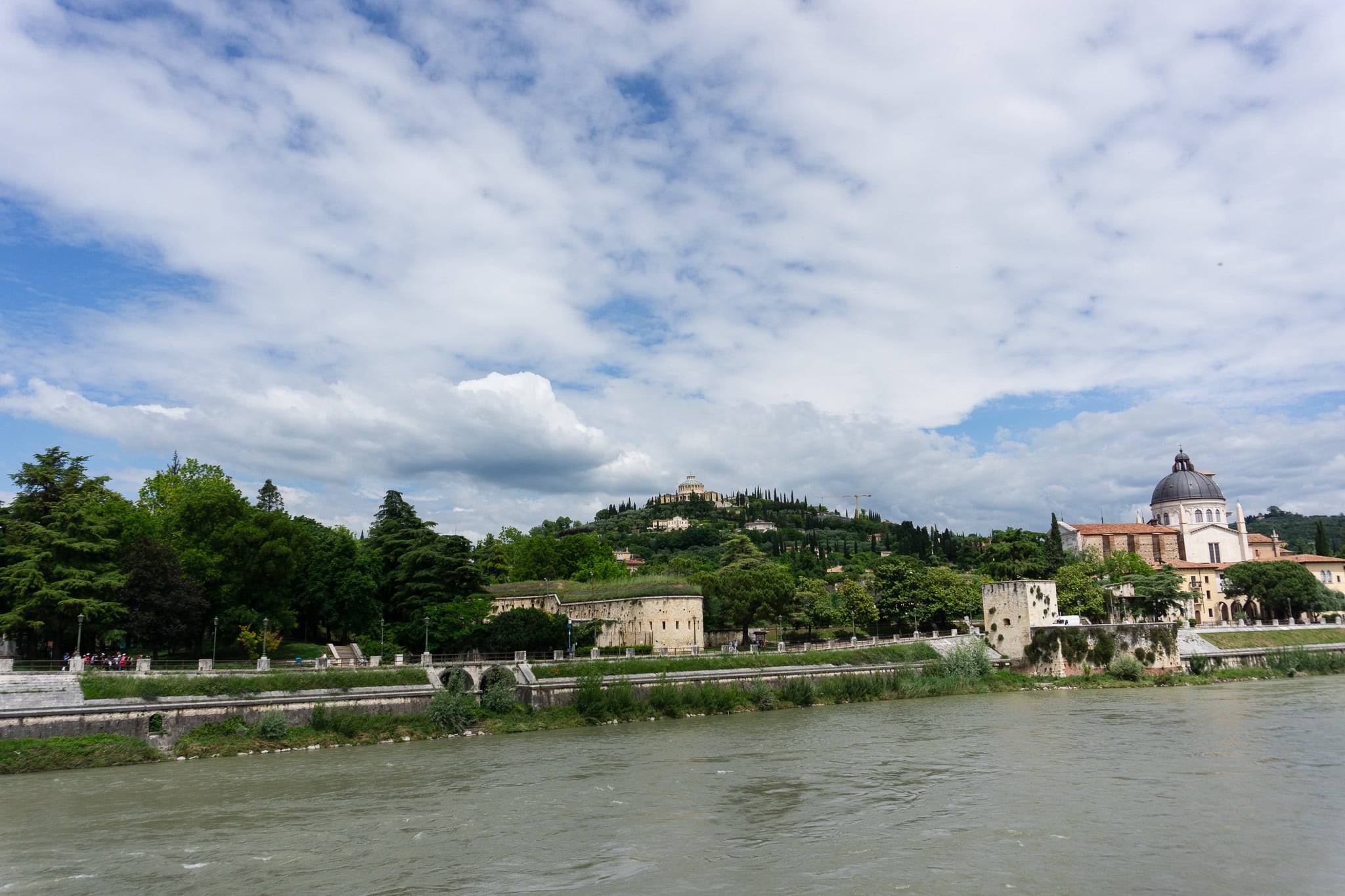 River view in Verona with stone embankment, domed church, green hillside, and people walking along the far bank.