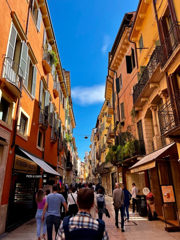 Pedestrian street in Verona lined with warm-toned historic buildings, bakery and gelato signs, and people walking beneath shutters and balconies.