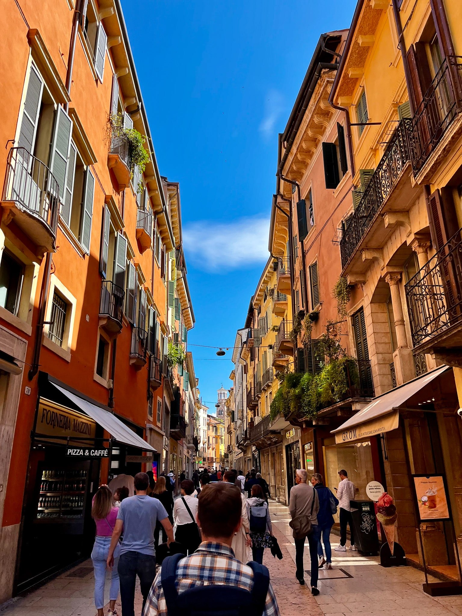 Pedestrian street in Verona lined with warm-toned historic buildings, bakery and gelato signs, and people walking beneath shutters and balconies.