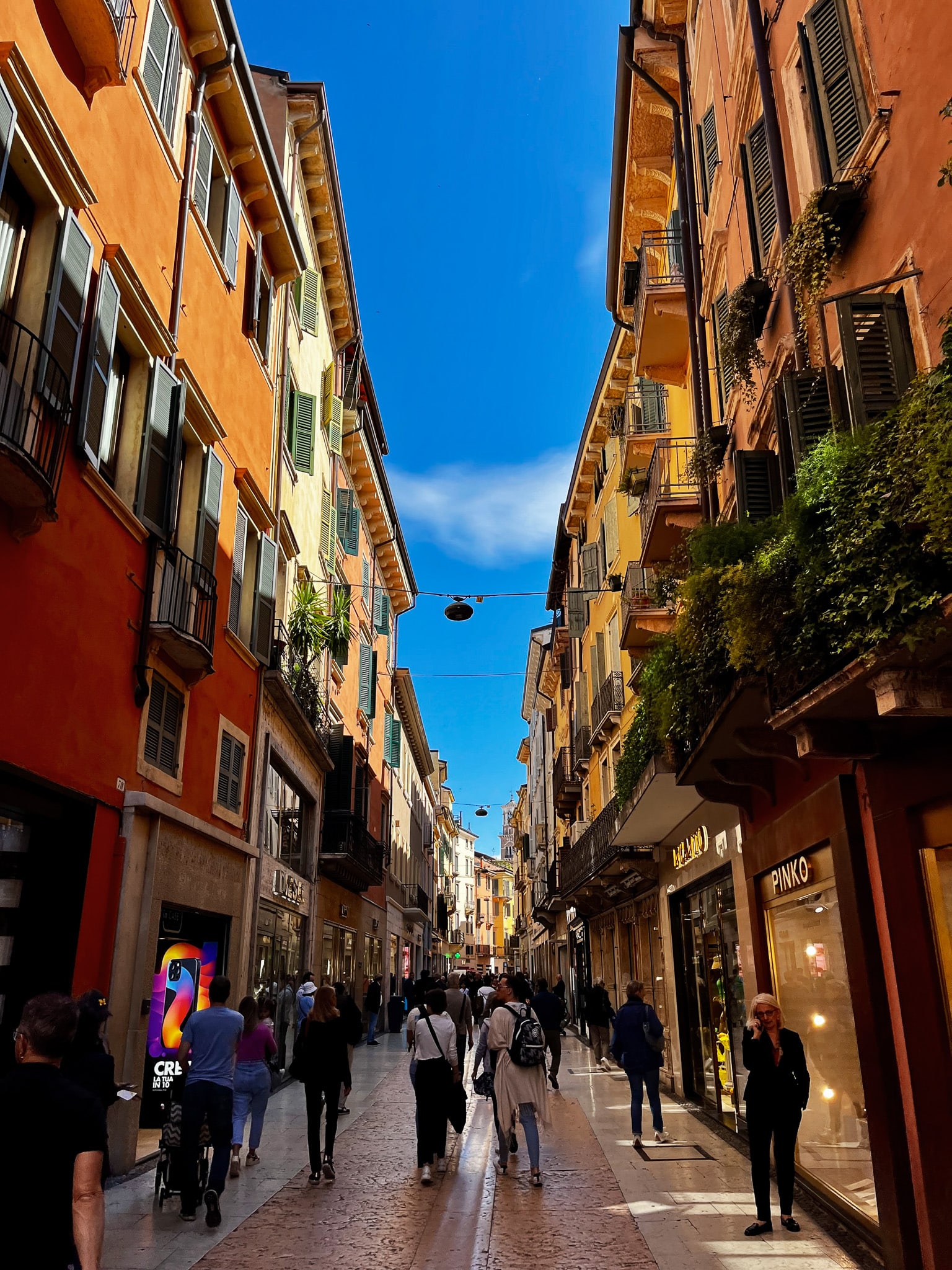 Narrow shopping street in Verona with tall pastel buildings, shutters, balconies, pedestrians, and a modern digital advertising display.