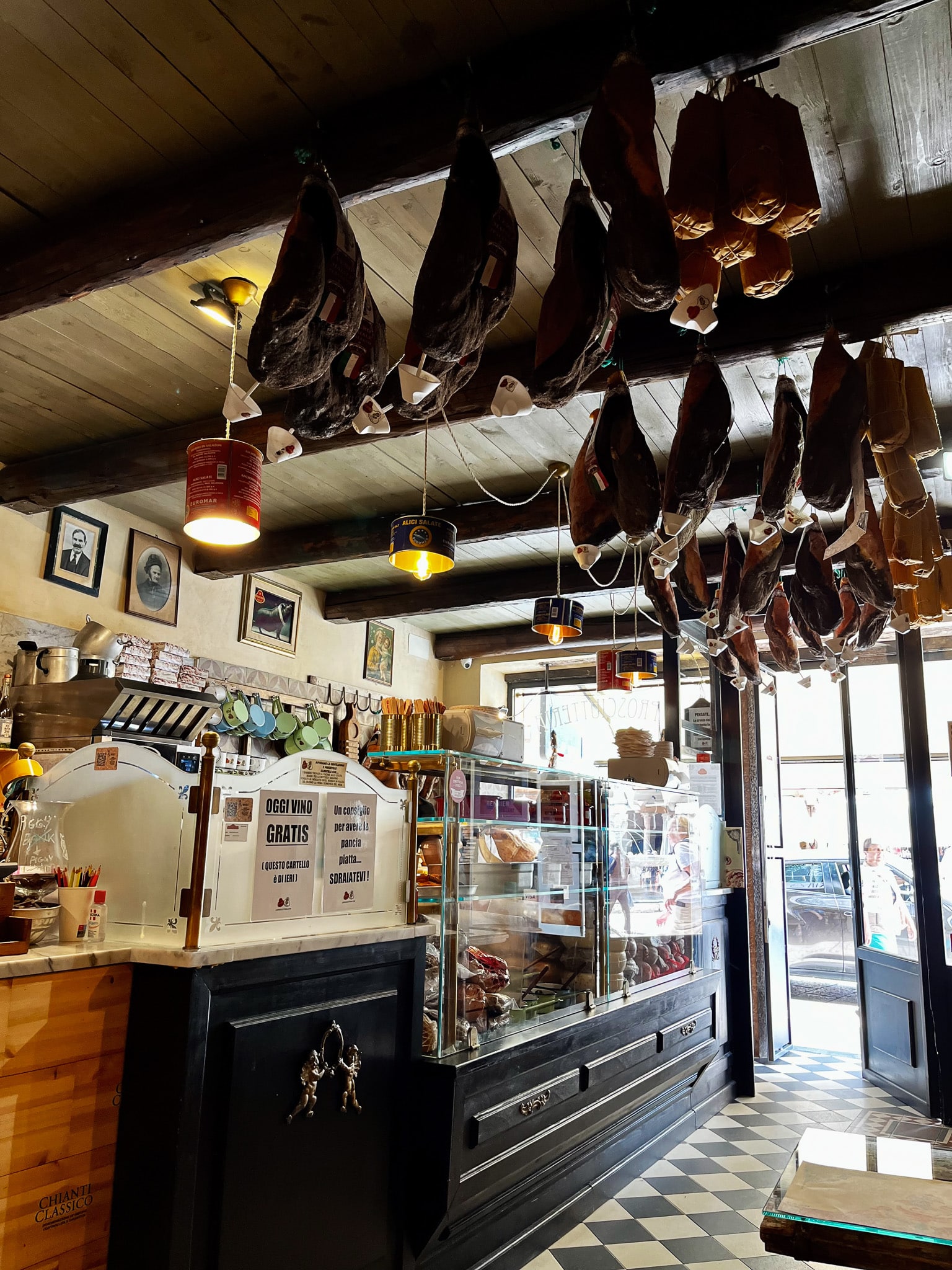 Interior of an Italian deli in Verona with hanging prosciutti, checkered floor, wooden beams, and product labels.