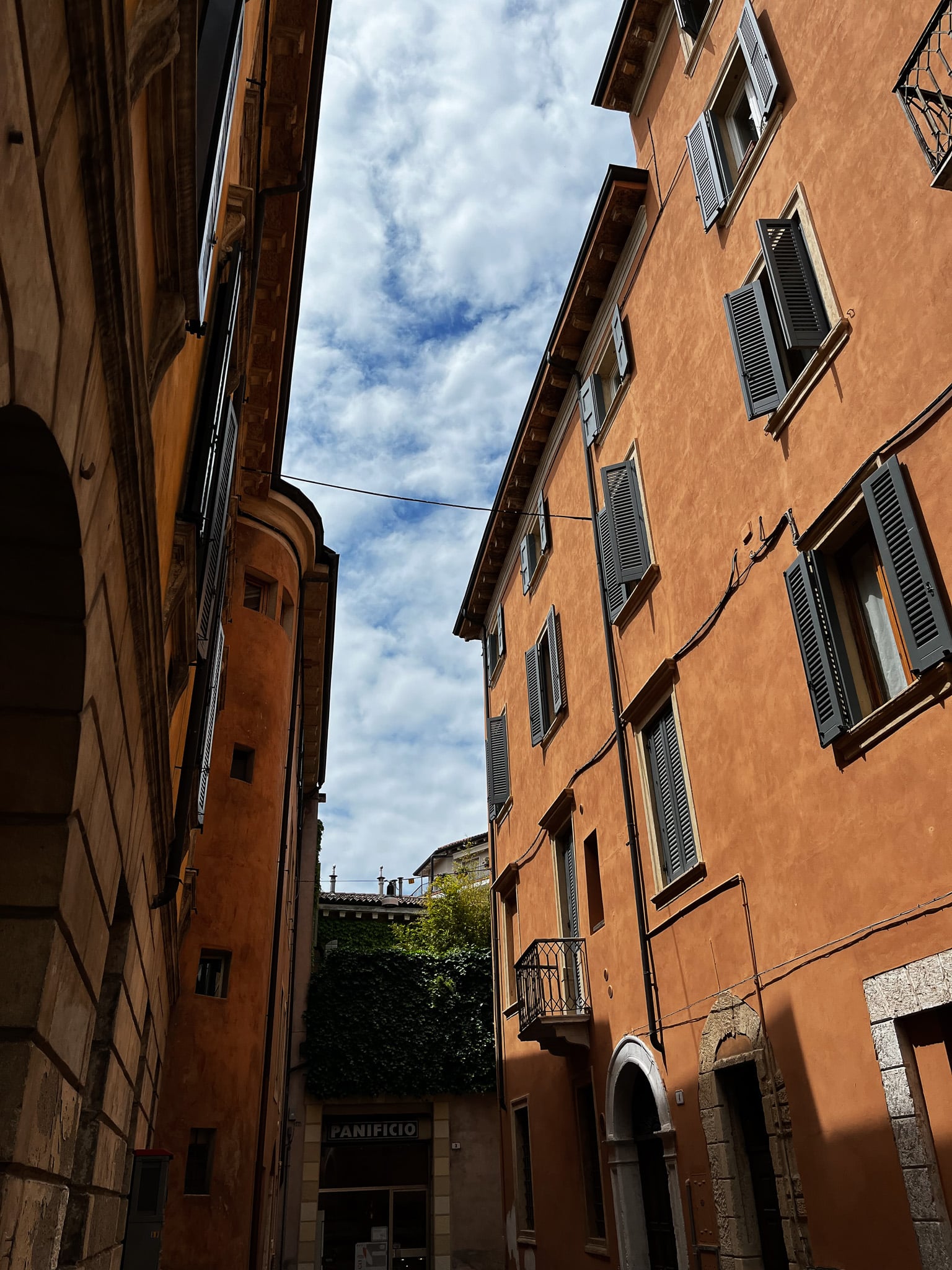 Upward view in a narrow Verona street with orange stucco walls, gray shutters, overhead wires, and a bakery sign reading PANIFICIO.