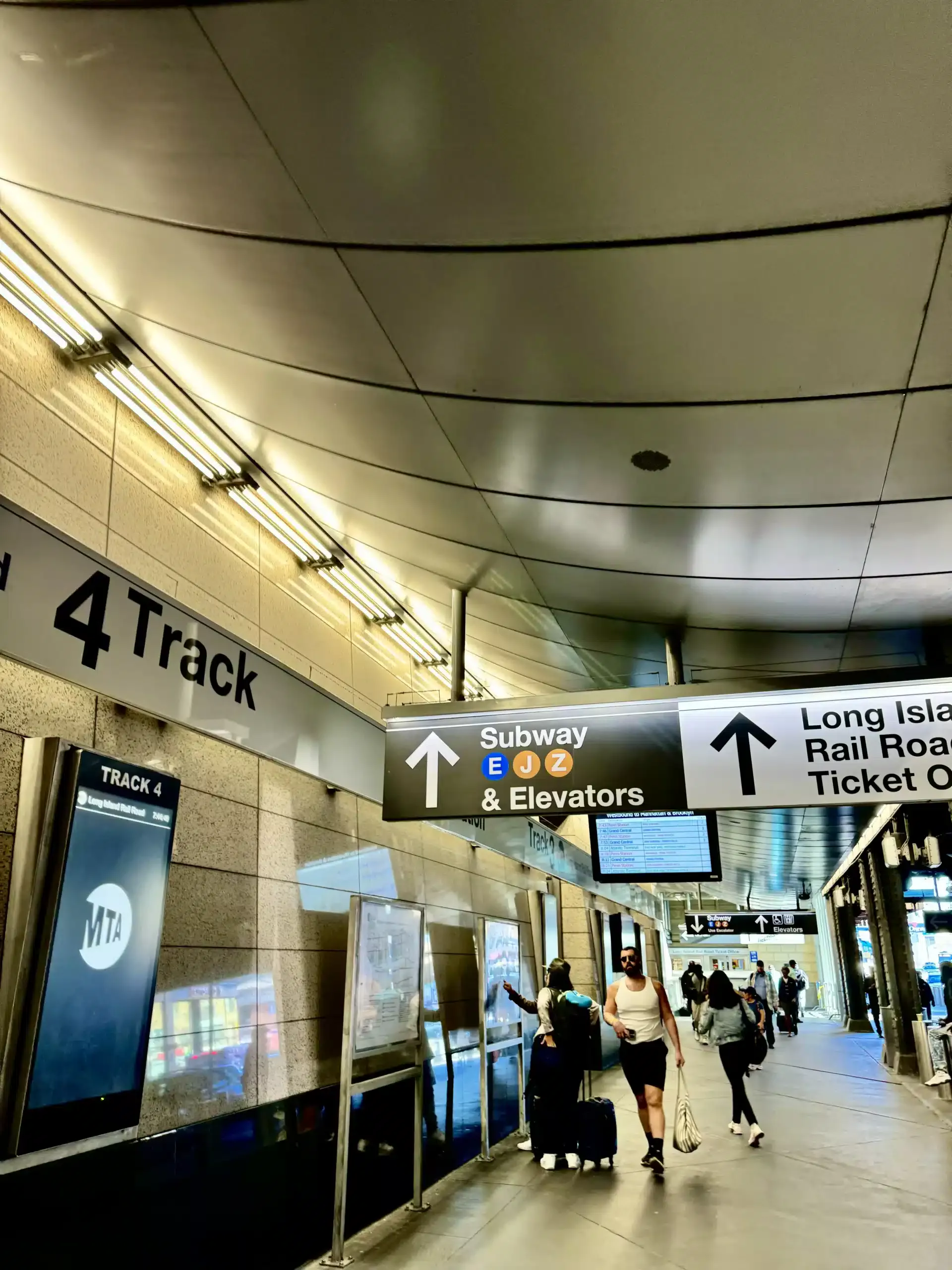 Transit concourse in New York with overhead wayfinding signs for Track 4, subway lines E J Z, elevators, and Long Island Rail Road connections.
