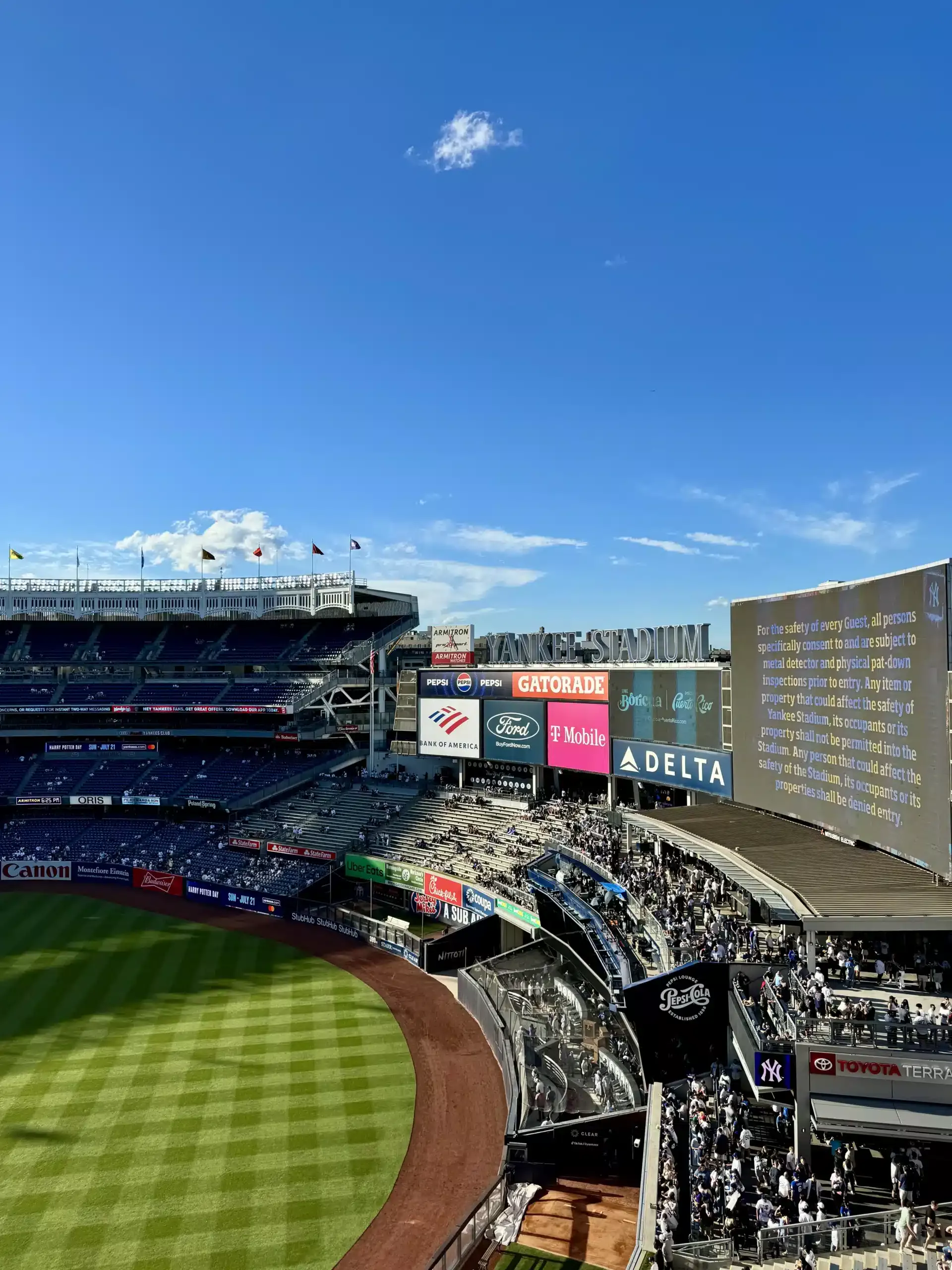 Wide interior view of Yankee Stadium with field, seating, sponsor signage, and a large guest safety notice board.