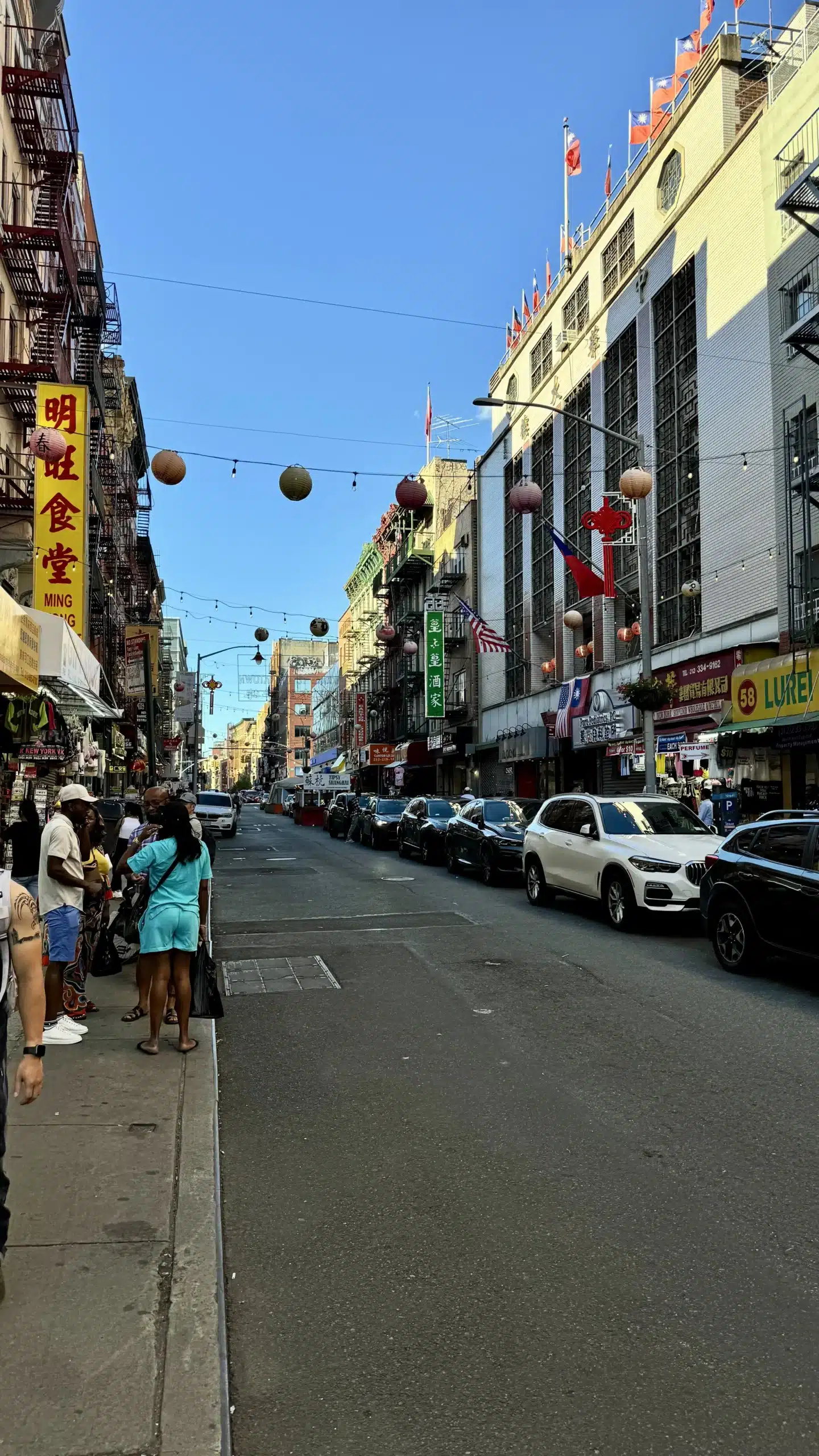 Chinatown street in New York with hanging lanterns, mixed-language storefront signs, flags, parked cars, and fire escapes.
