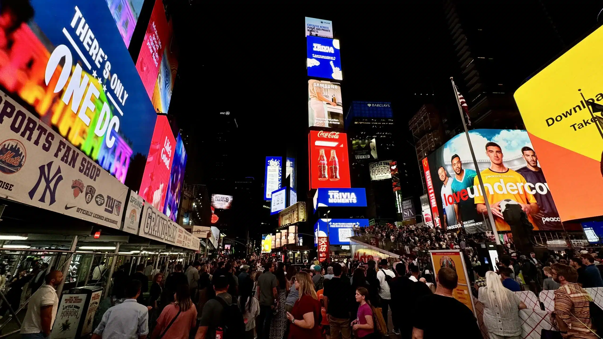 Night view of Times Square packed with crowds, digital billboards, Broadway ads, sports store signage, and stepped seating.