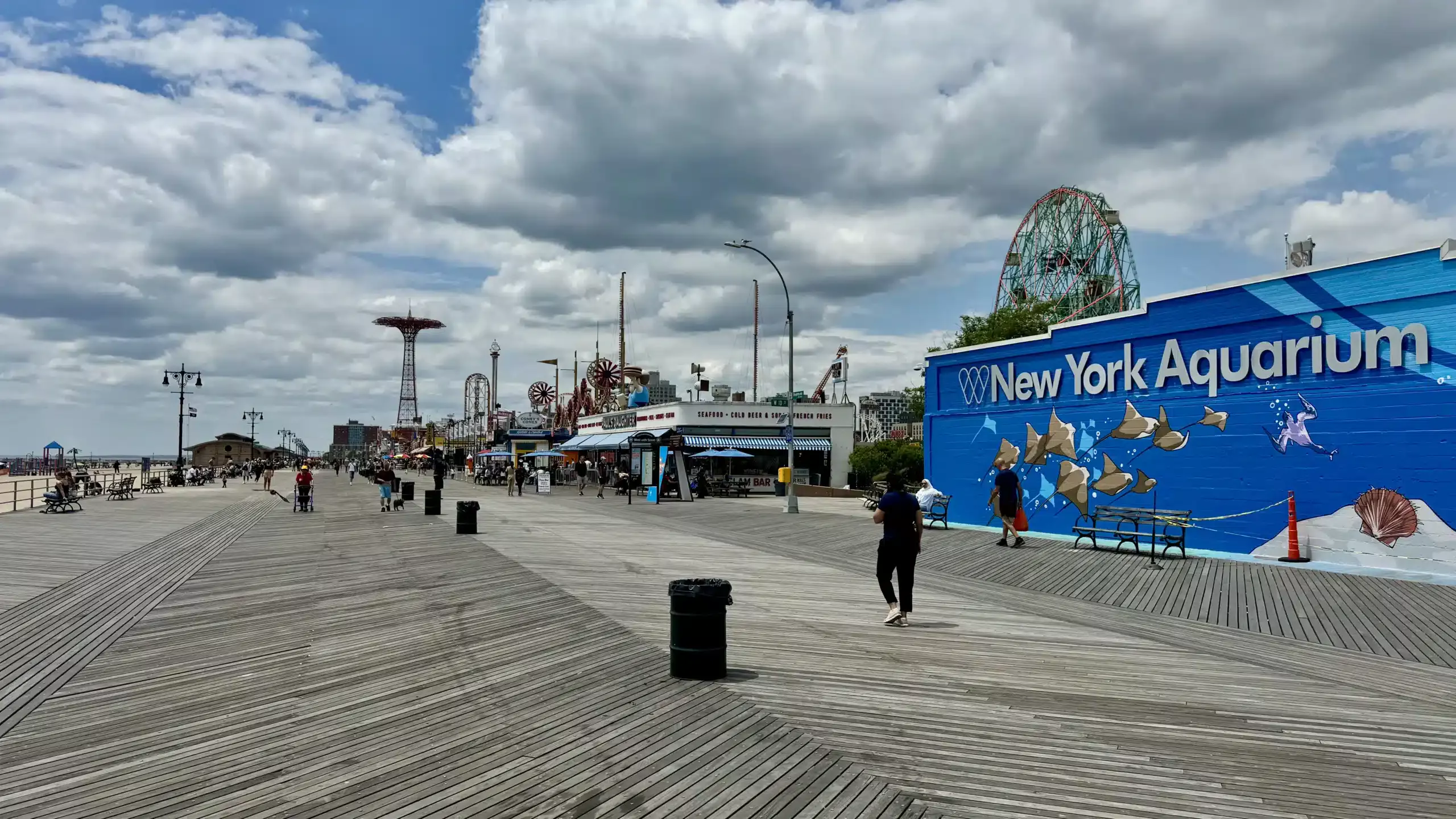 Coney Island boardwalk with large New York Aquarium mural, food stand signs, benches, pedestrians, and amusement rides in the distance.