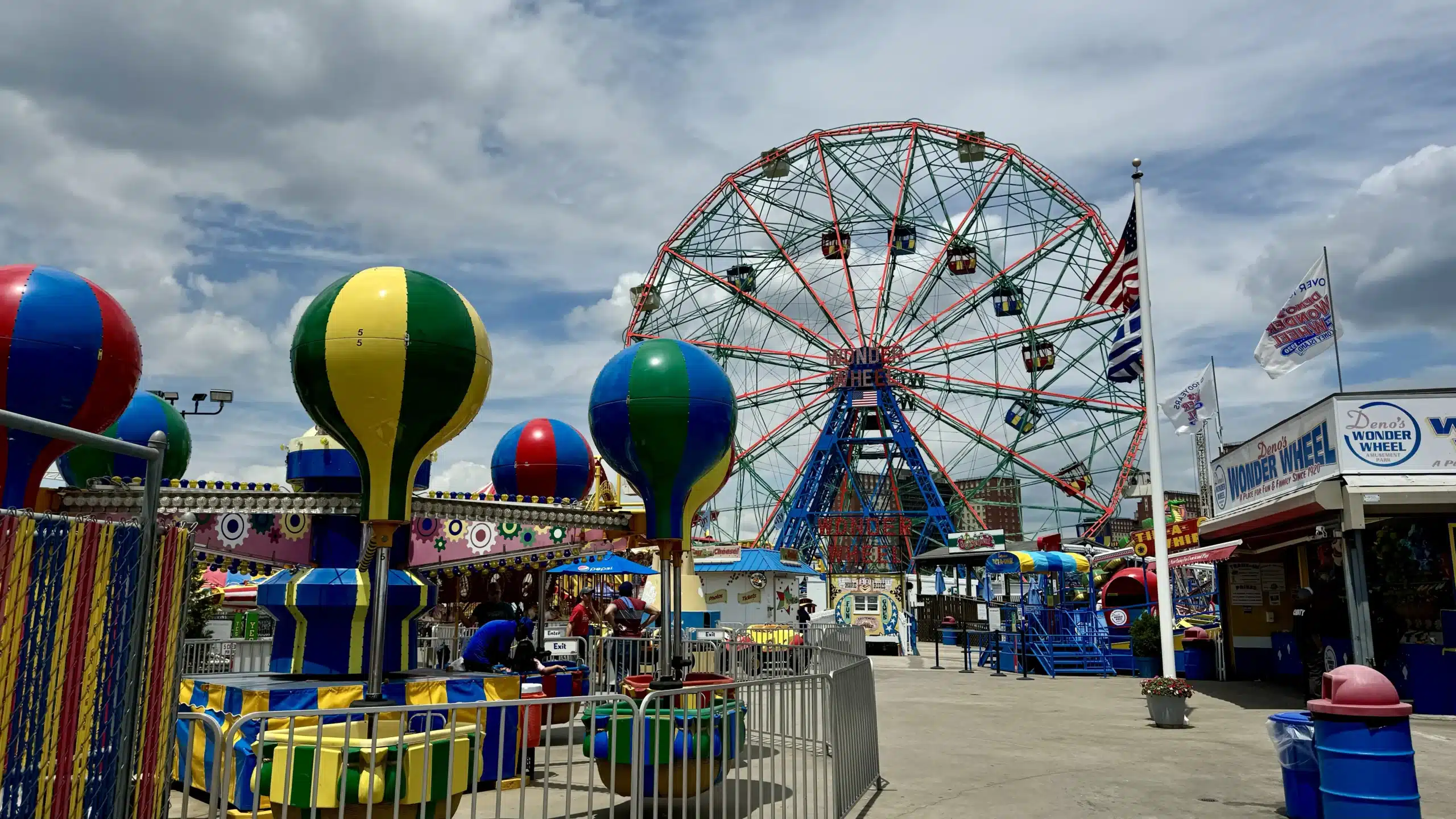 Coney Island amusement area with Deno's Wonder Wheel, entry and exit signs, ticket booths, queue barriers, and colorful rides.