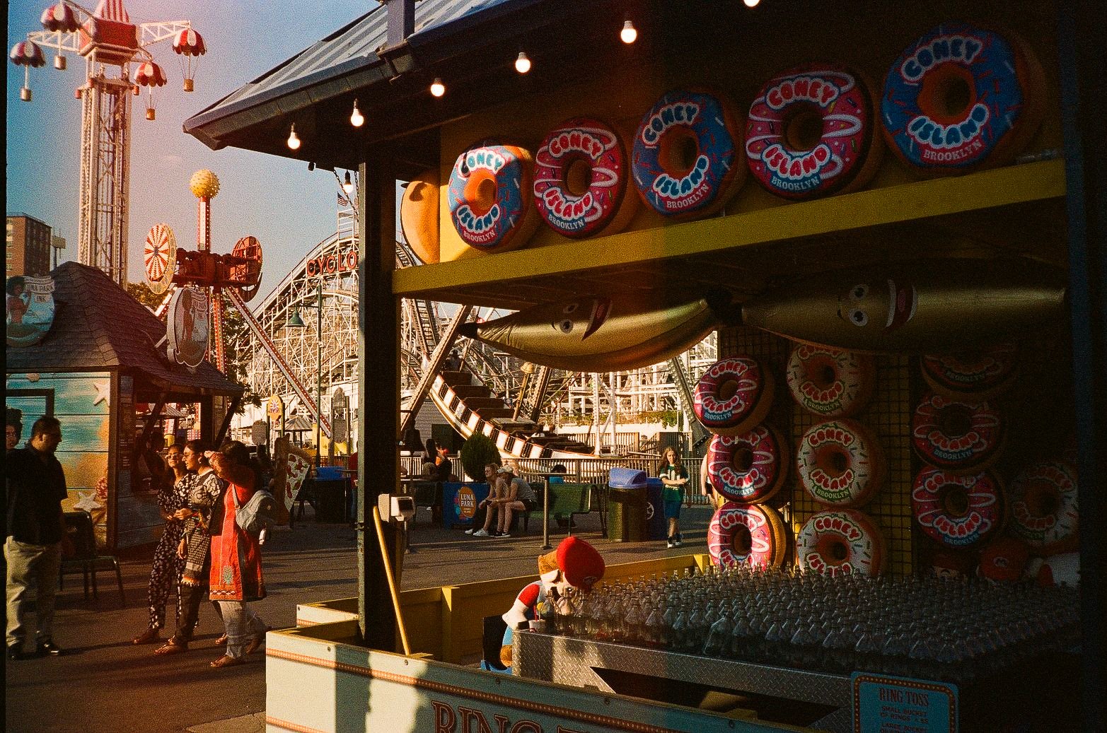 Coney Island and Brooklyn signs at a midway stall with roller coaster structure and Luna Park visible behind.