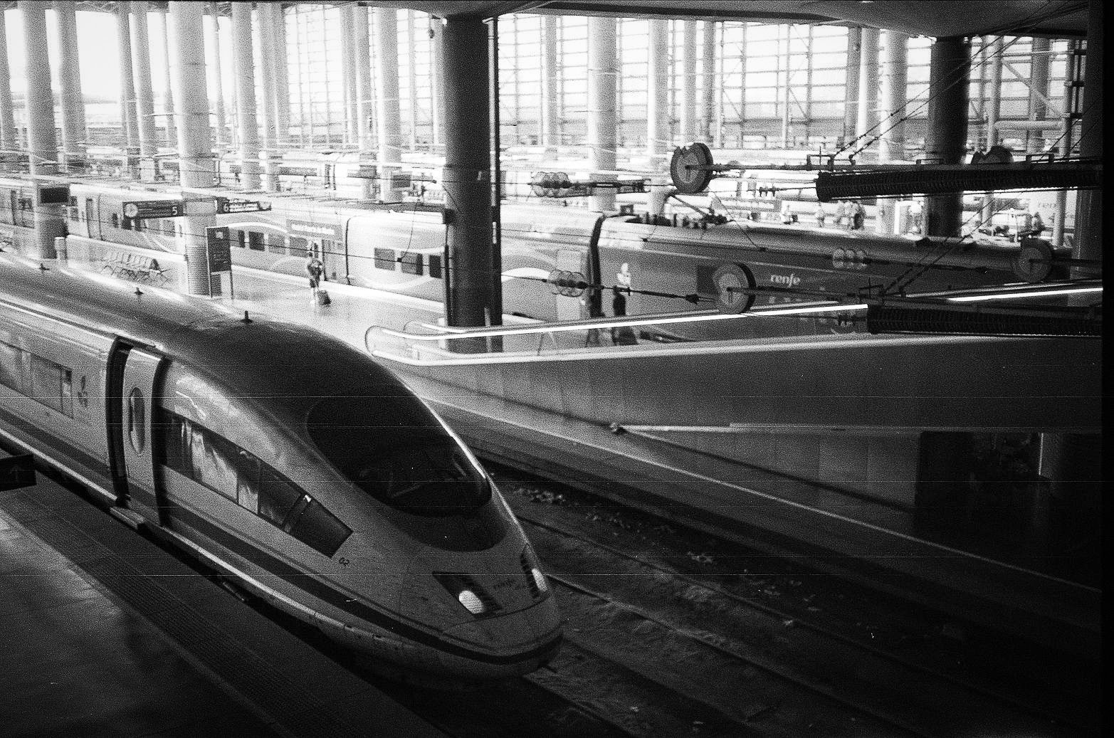 Indoor rail station with streamlined high-speed trains at platforms beneath a large glass-and-steel roof structure.