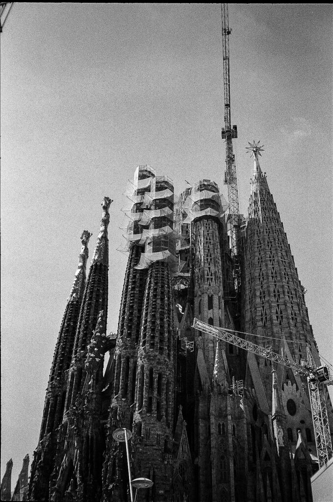 Low-angle black-and-white view of the Sagrada Família with ornate spires, scaffolding, cranes, and the star-topped tower visible above.