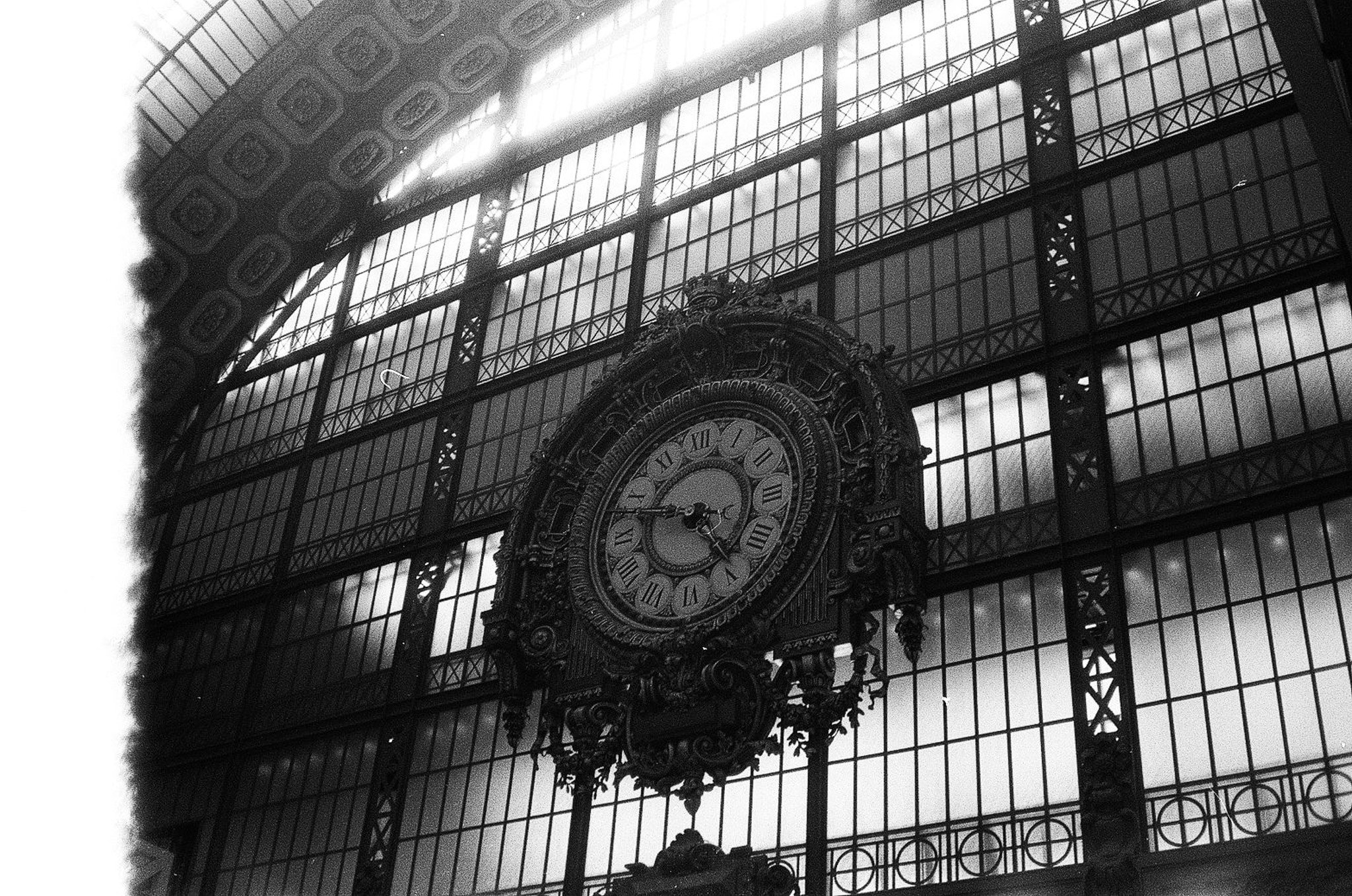 Black-and-white interior with an ornate giant clock, iron framing, and a tall glass roof in a historic Paris hall, likely the Musée d’Orsay.