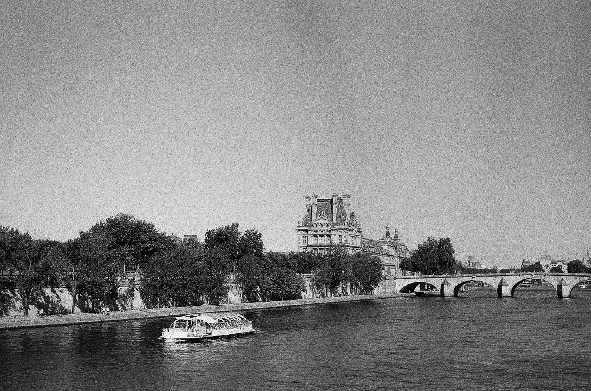 Black-and-white Seine river scene with a stone bridge, passenger boat, tree-lined embankment, and historic building in Paris.