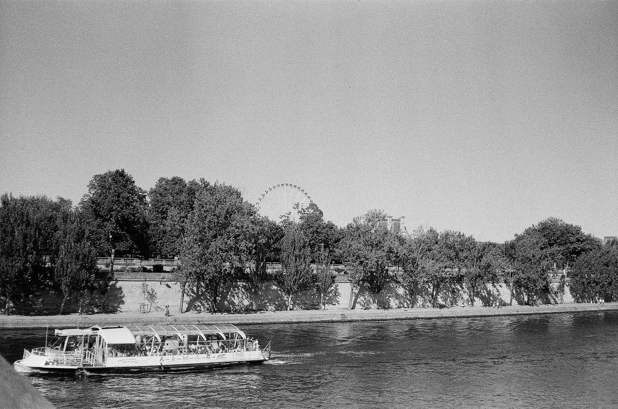 Black-and-white riverfront view with a passenger boat on the Seine, tree-lined embankment, and Ferris wheel in the background.