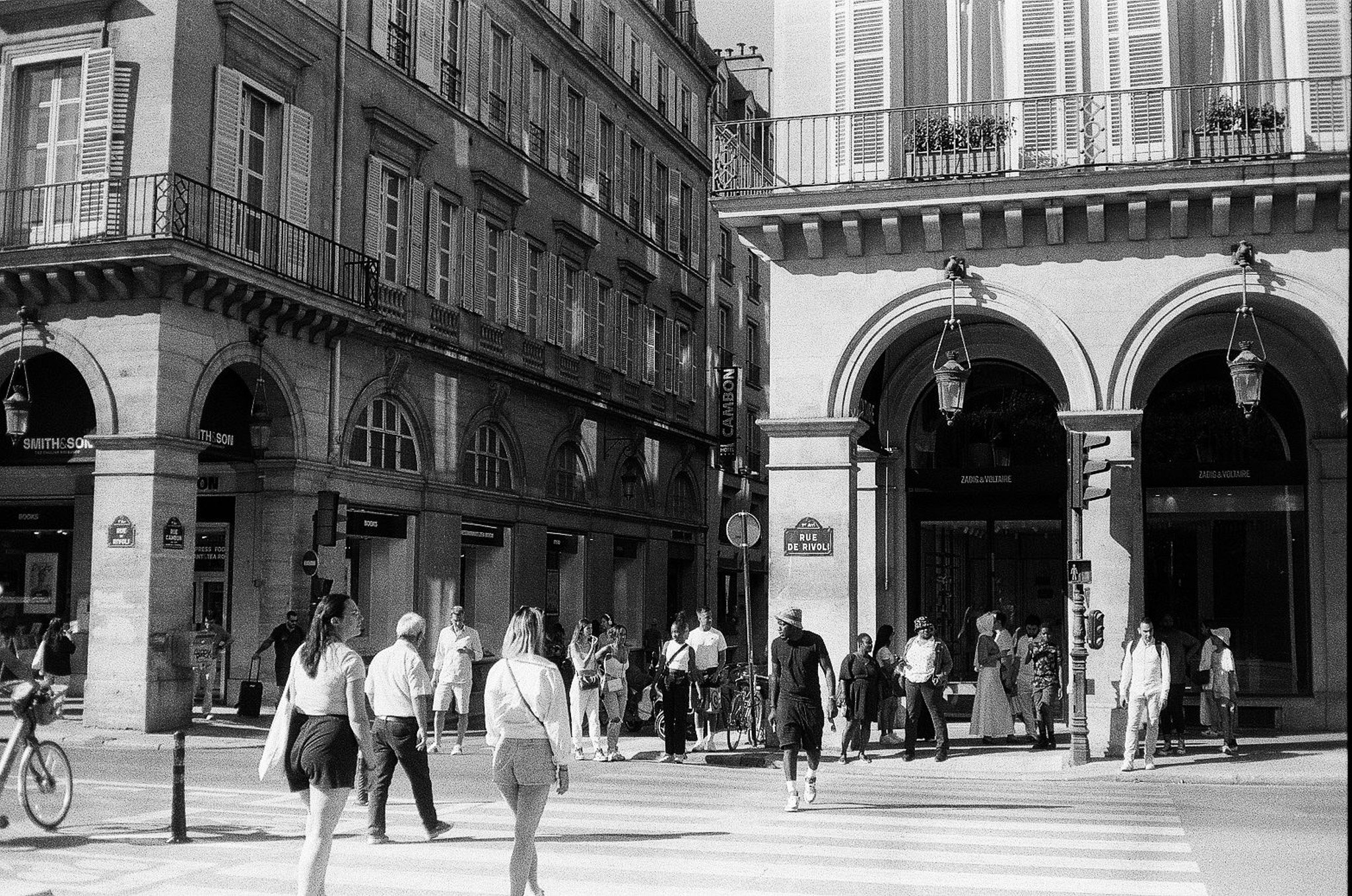Black-and-white street corner on Rue de Rivoli with arcaded buildings, pedestrians crossing, retail signs, and historic facades in Paris.