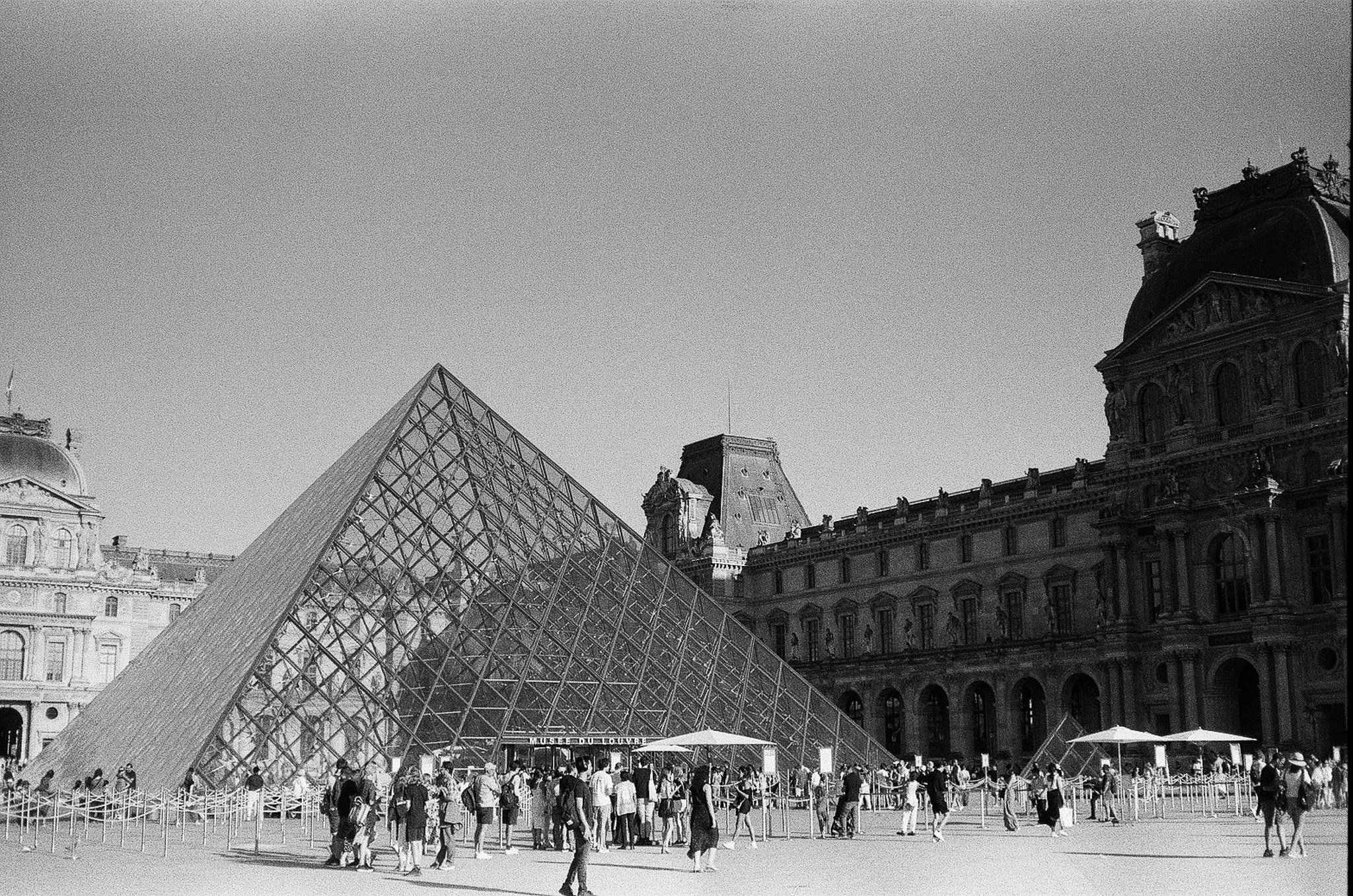 Black-and-white view of the Louvre Pyramid with queue barriers, canopies, crowds, and historic palace buildings around it in Paris.