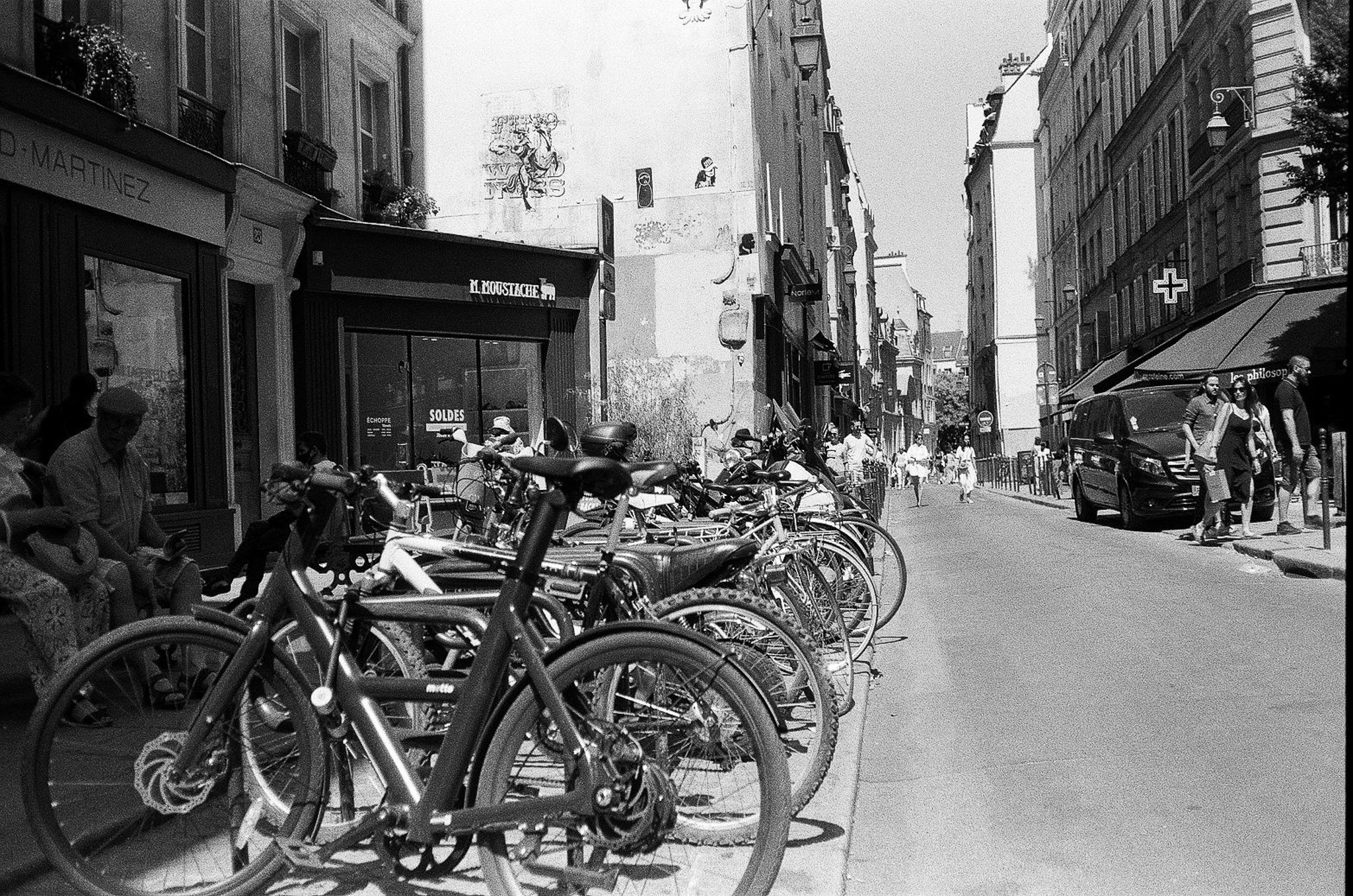 Black-and-white narrow Paris street with many parked bicycles, pedestrians, storefronts, and sale signage.