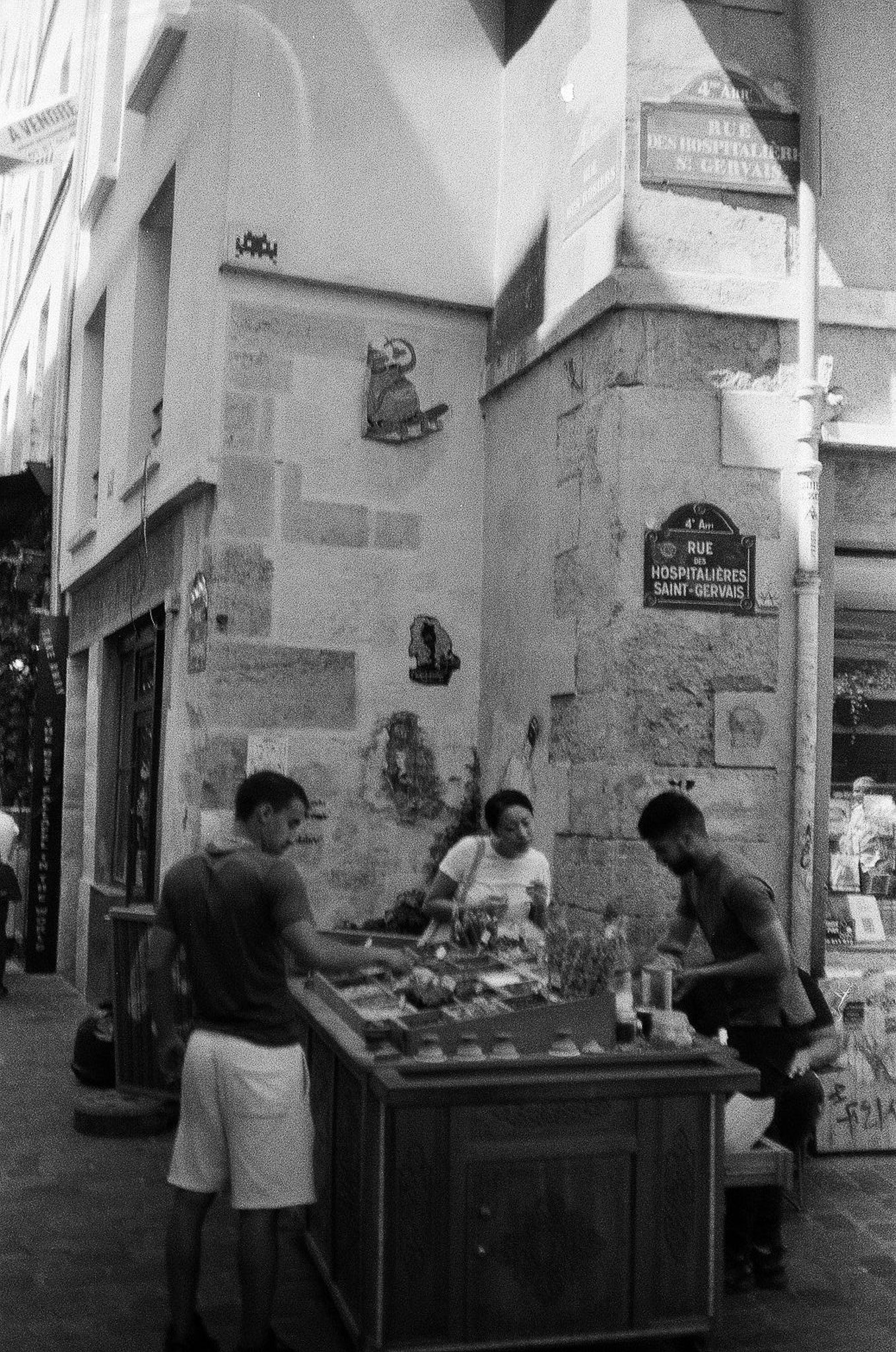 Black-and-white corner scene on Rue des Hospitalières-Saint-Gervais in Paris 4th arrondissement with old facade, street sign, and people near a small street table.