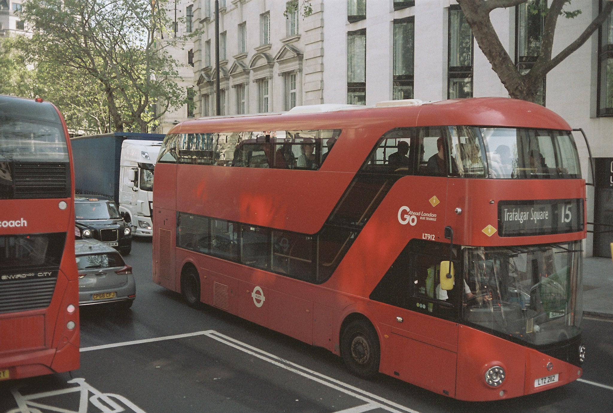 Red London bus with destination display reading Trafalgar Square 15 on a busy tree-lined street.