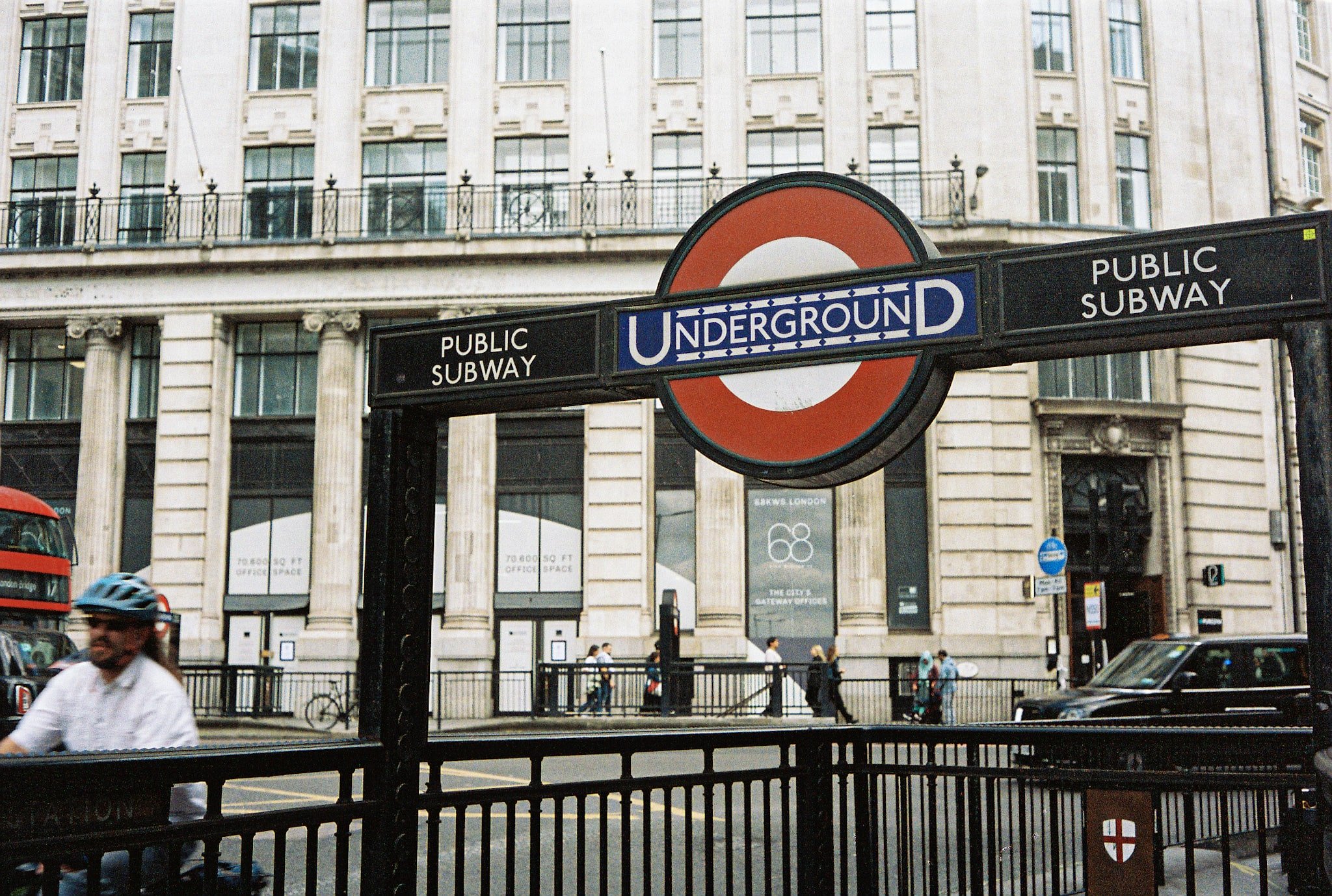 London Underground entrance with roundel sign, public subway panels, railings, pedestrians and a stone building across the street.