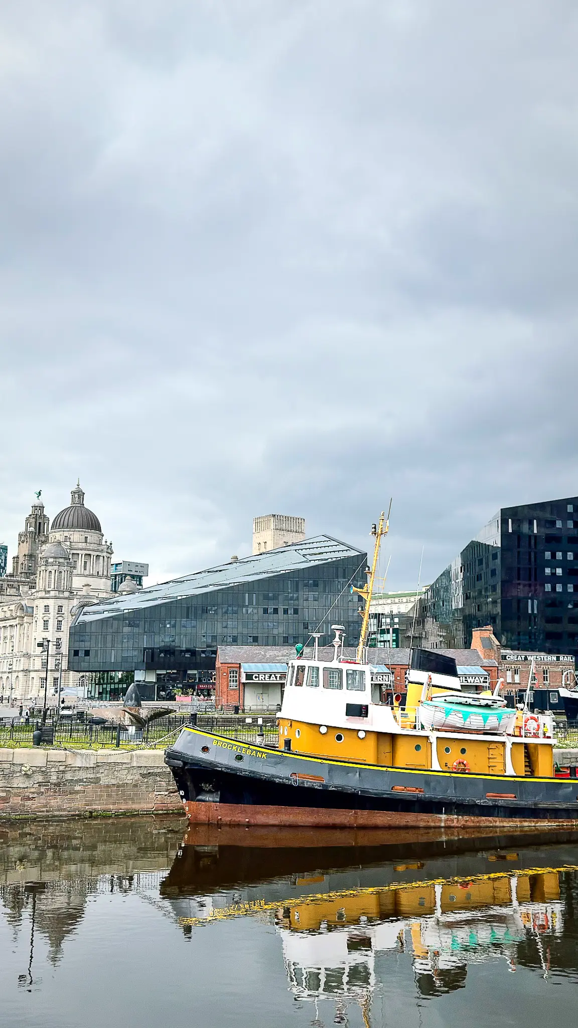 Brocklebank tugboat moored on Liverpool waterfront with historic buildings and dark angular Mann Island development behind.