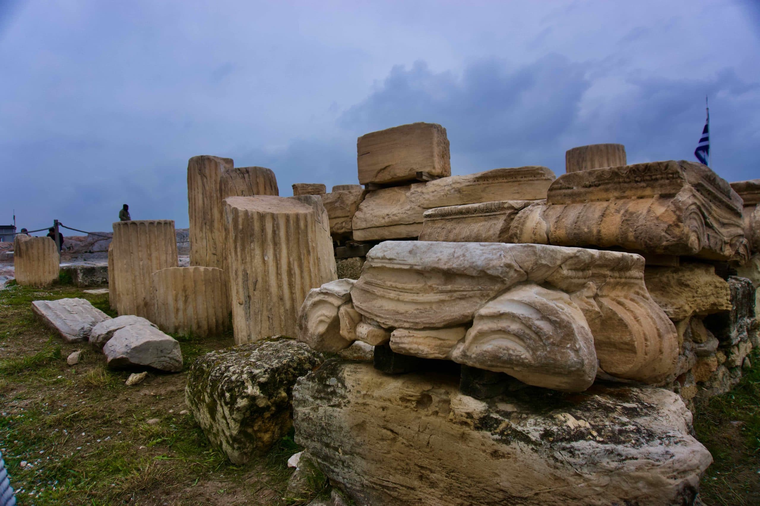 Scattered ancient stone fragments at an archaeological site in Greece with Greek flag