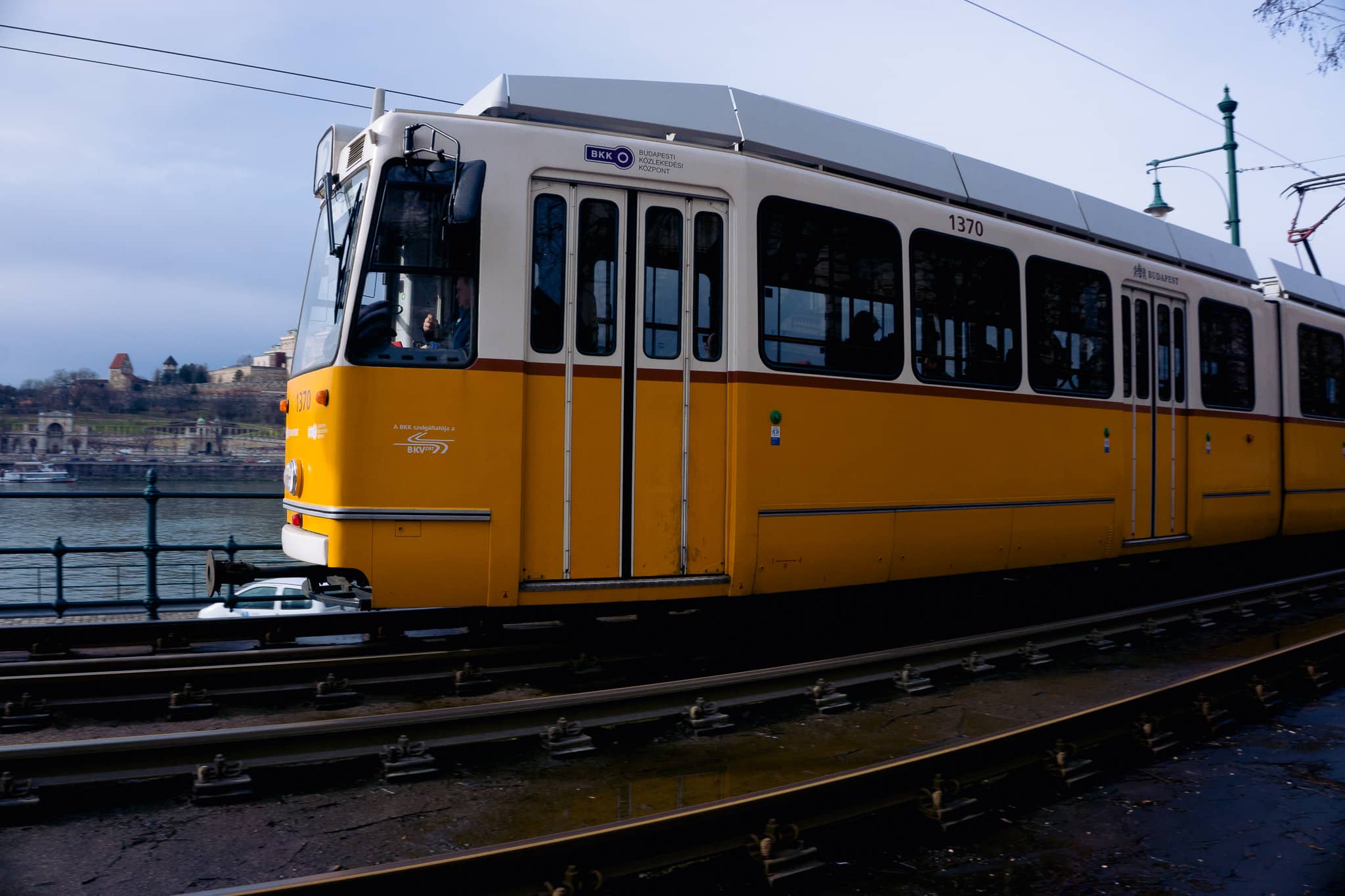 Side view of a Budapest tram with BKK branding traveling beside wet tram tracks near the river
