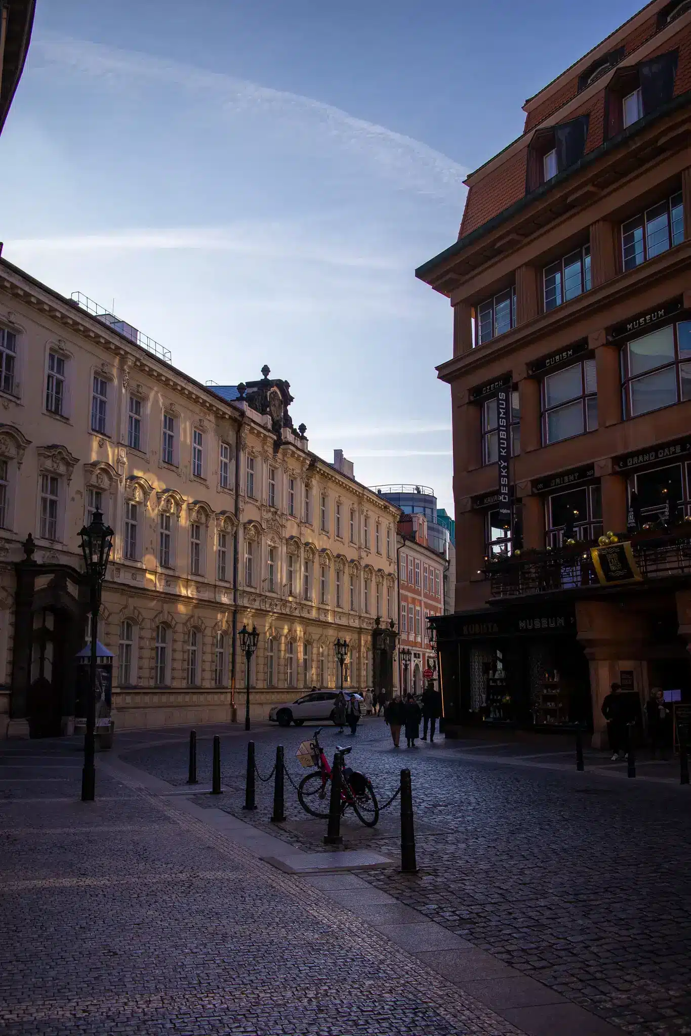 Historic Prague street with museum signage for Czech Cubism, cafés, cobblestones, and a parked bicycle.