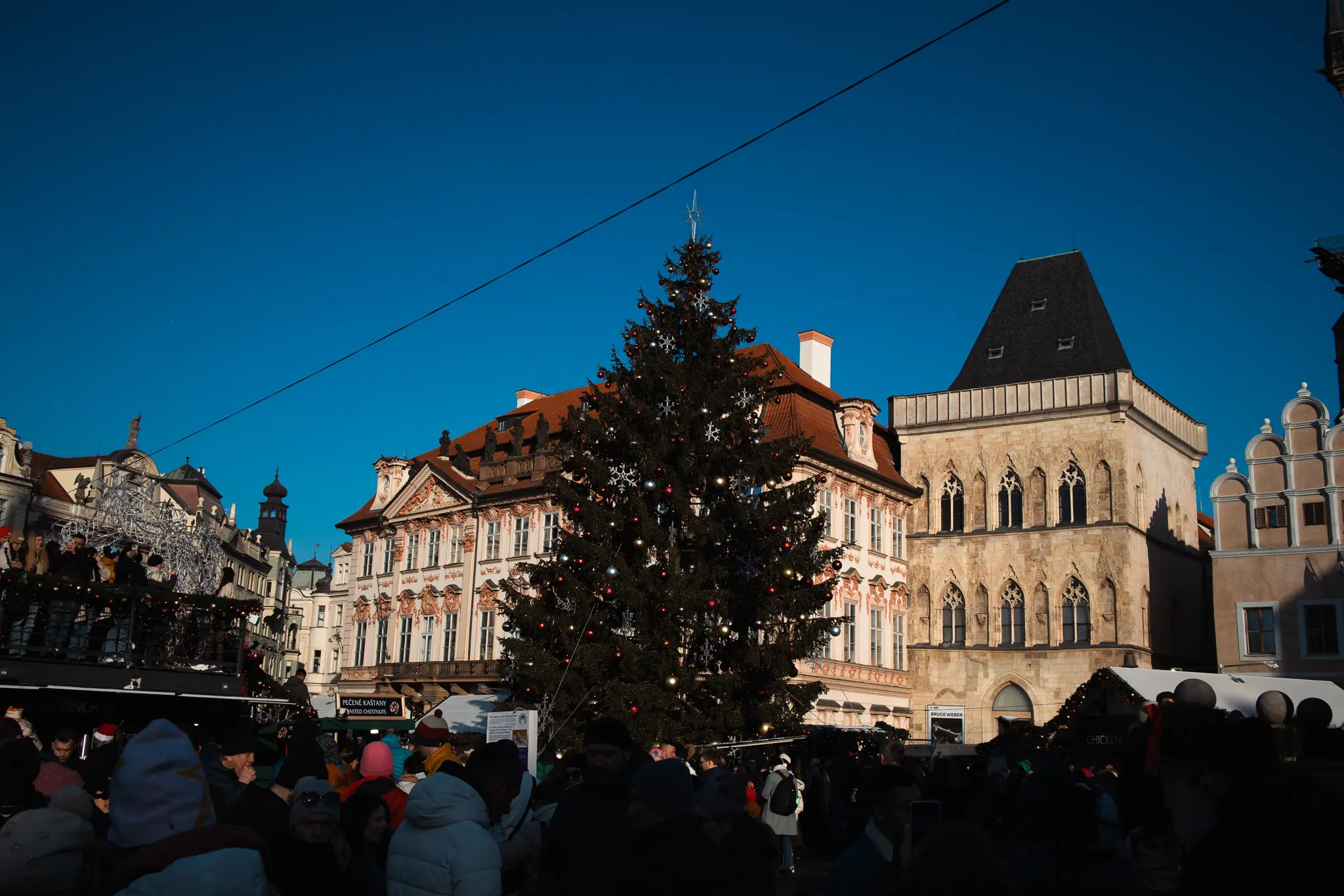 Christmas tree and roasted chestnut stall in a crowded historic square in Prague under clear winter daylight.