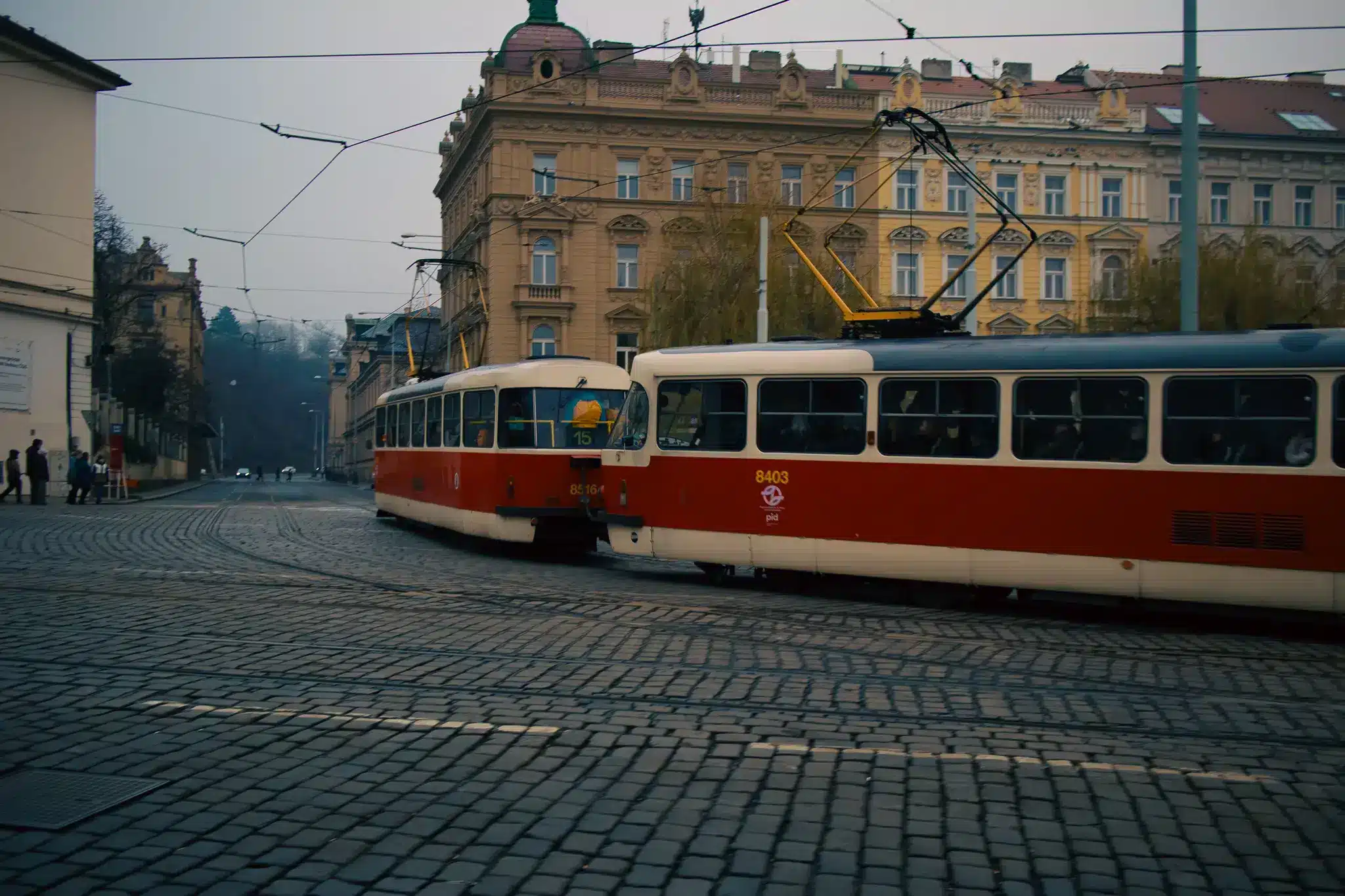 Two Prague trams crossing a cobblestone intersection beneath overhead wires in front of an ornate historic building.