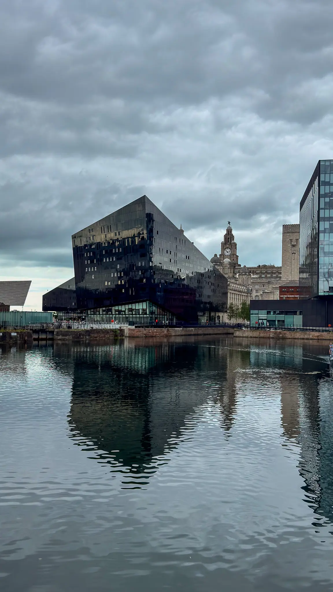 Dark angular Mann Island building beside Liverpool waterfront with older clock tower and modern glass buildings under cloudy sky.