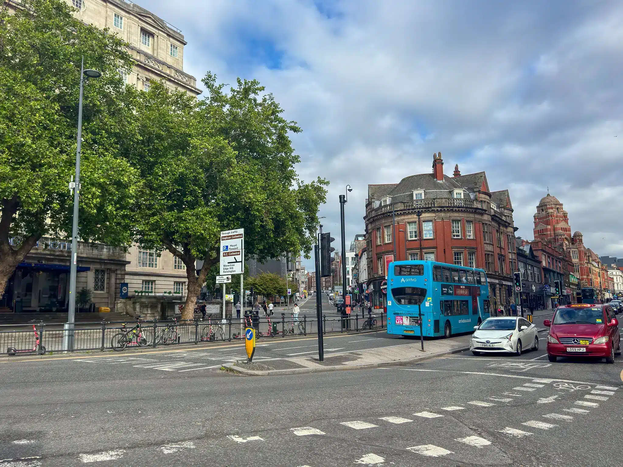 Busy Liverpool road junction with signs pointing to Anglican Cathedral and R.C. Cathedral, bus and historic buildings.