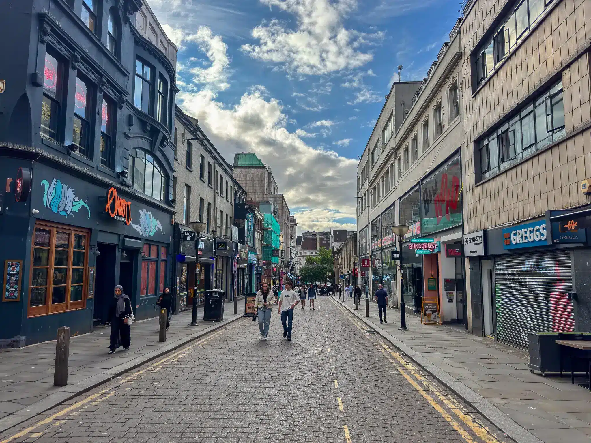 Pedestrian street in Liverpool with Greggs, Bundobust and other storefronts along cobbled paving.