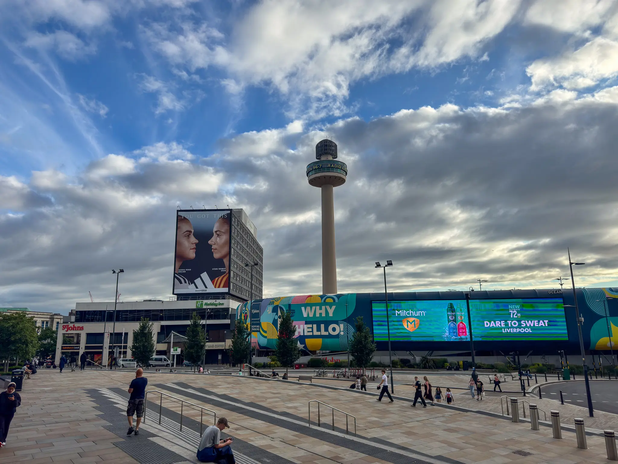 Wide Liverpool plaza with RADIO CITY tower, St Johns signage, billboard screens and scattered pedestrians.