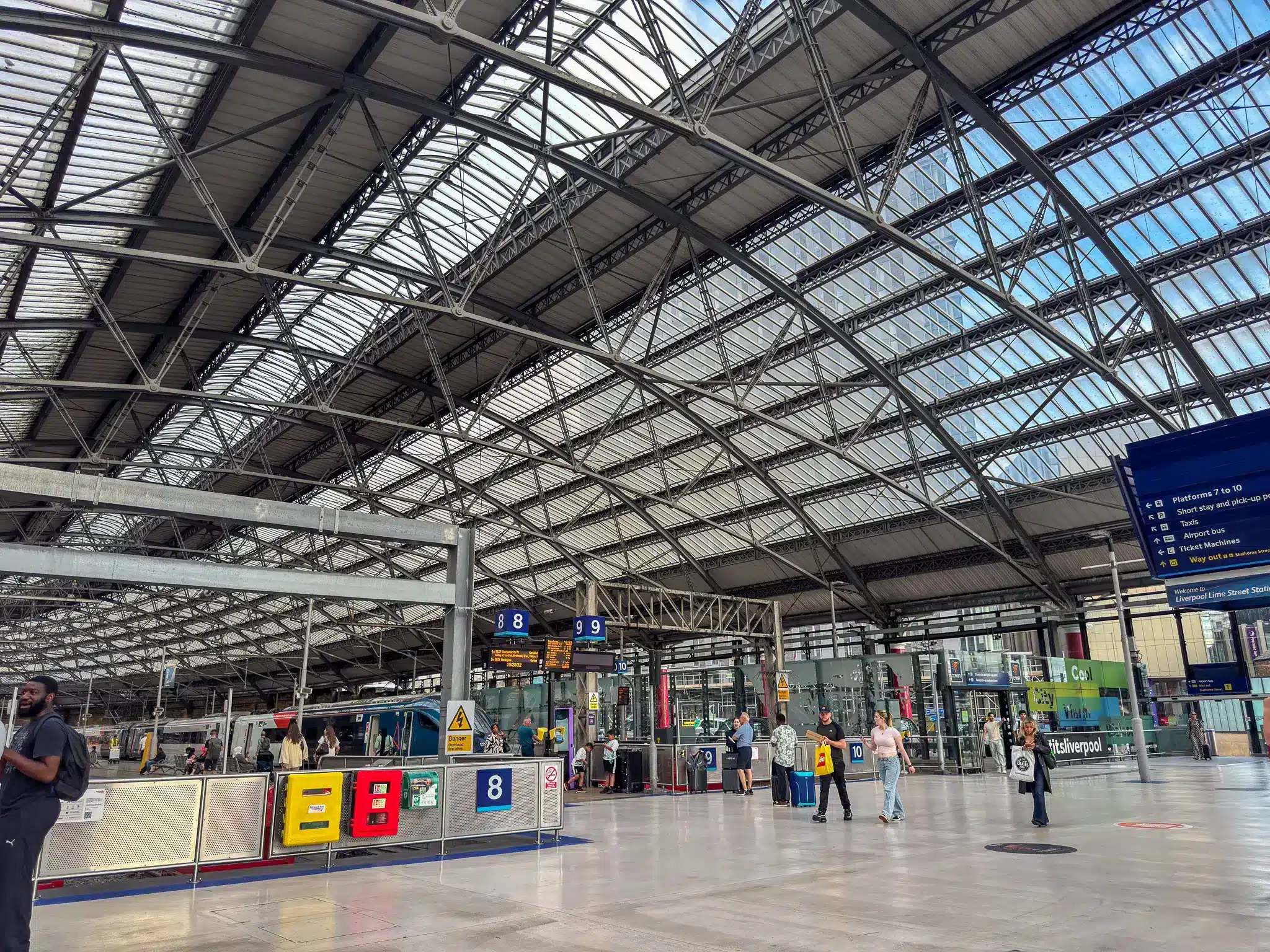 Interior of Liverpool Lime Street Station with platforms, arched roof, passengers and signs for taxis and airport bus.